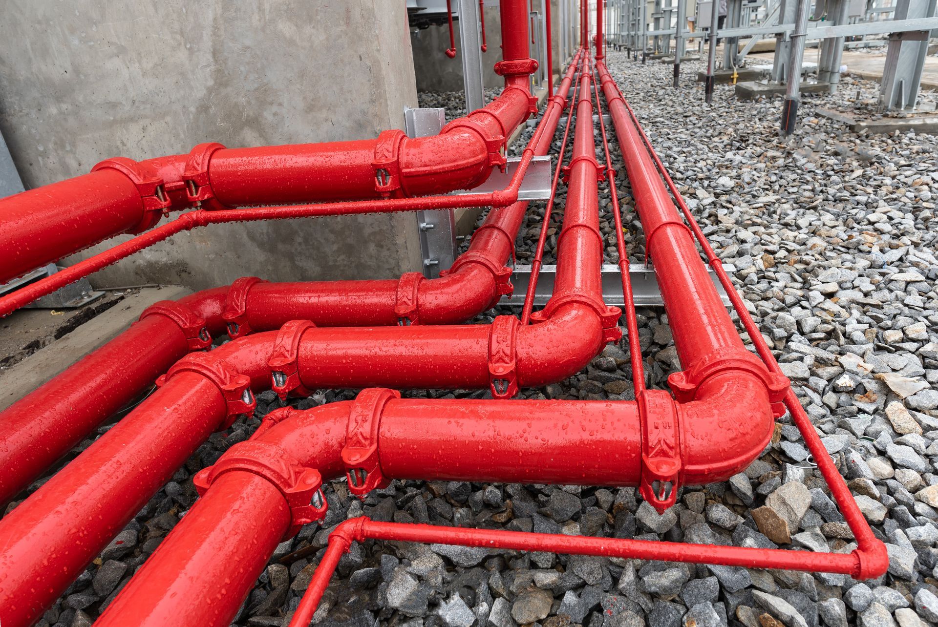 Red fire sprinkler pipes outside, against a concrete wall and gravel.