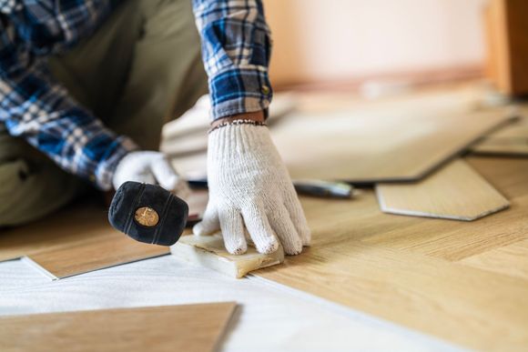 Person in plaid shirt and gloves installing light wood flooring.