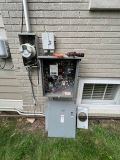 Open electrical panel on a brick building exterior, tools visible, grass below.