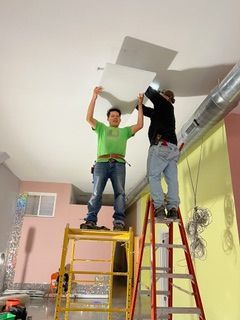 Electrician kneeling, testing wires near wall outlets with a multimeter. Toolbox open.