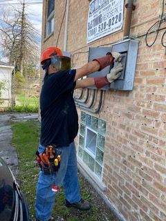 Electrician in orange hard hat, gloves, and safety glasses working on electrical panel outside on brick wall.