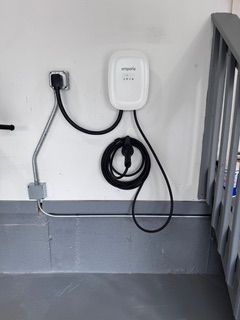 Electric vehicle charger mounted on a white wall near stairs. Black cable looped below charger.