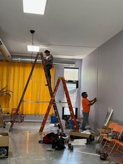 Two workers installing electrical wiring in a room. One on a ladder, one on the wall.