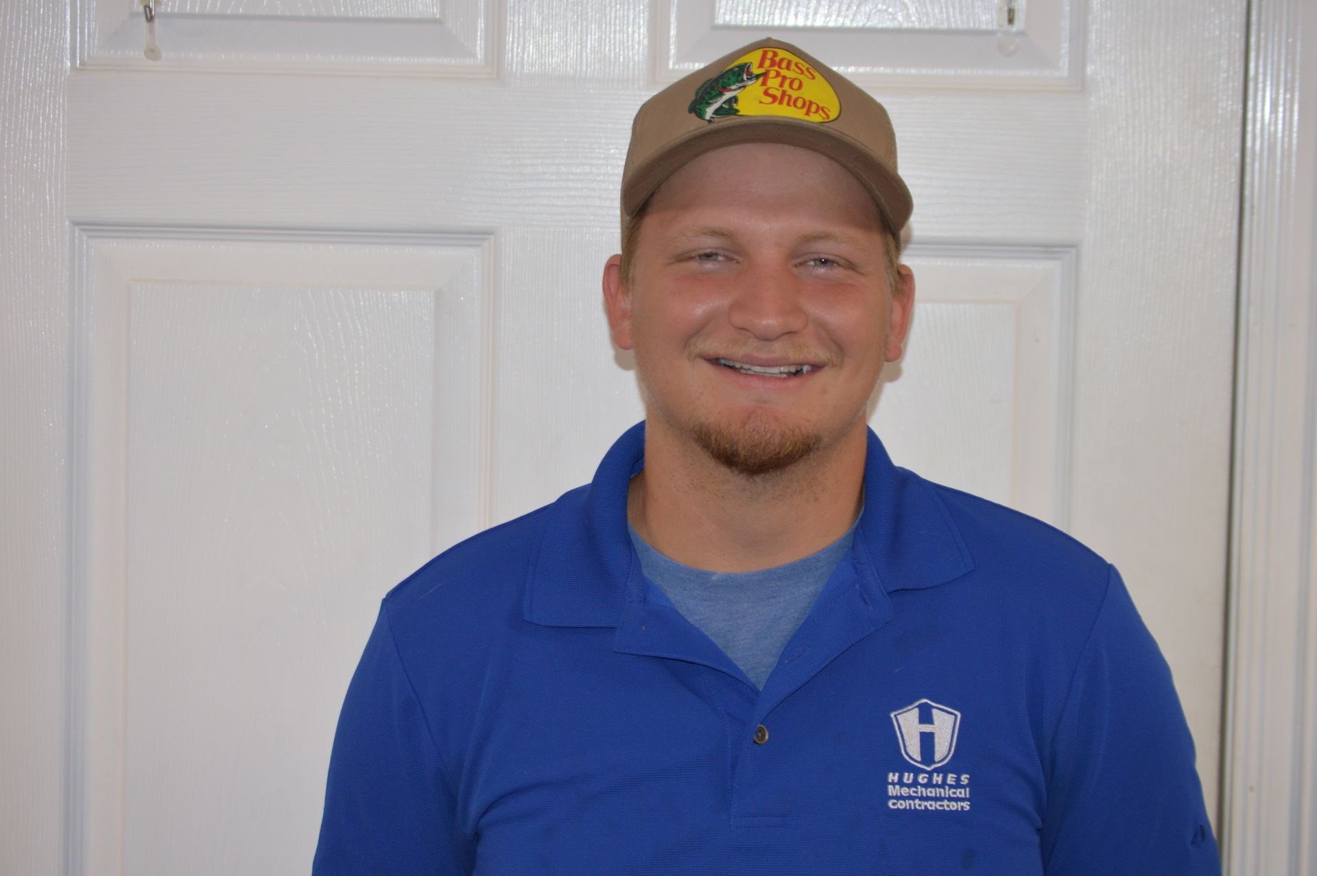 Man wearing a Bass Pro Shops cap and blue polo shirt smiles in front of a white door.
