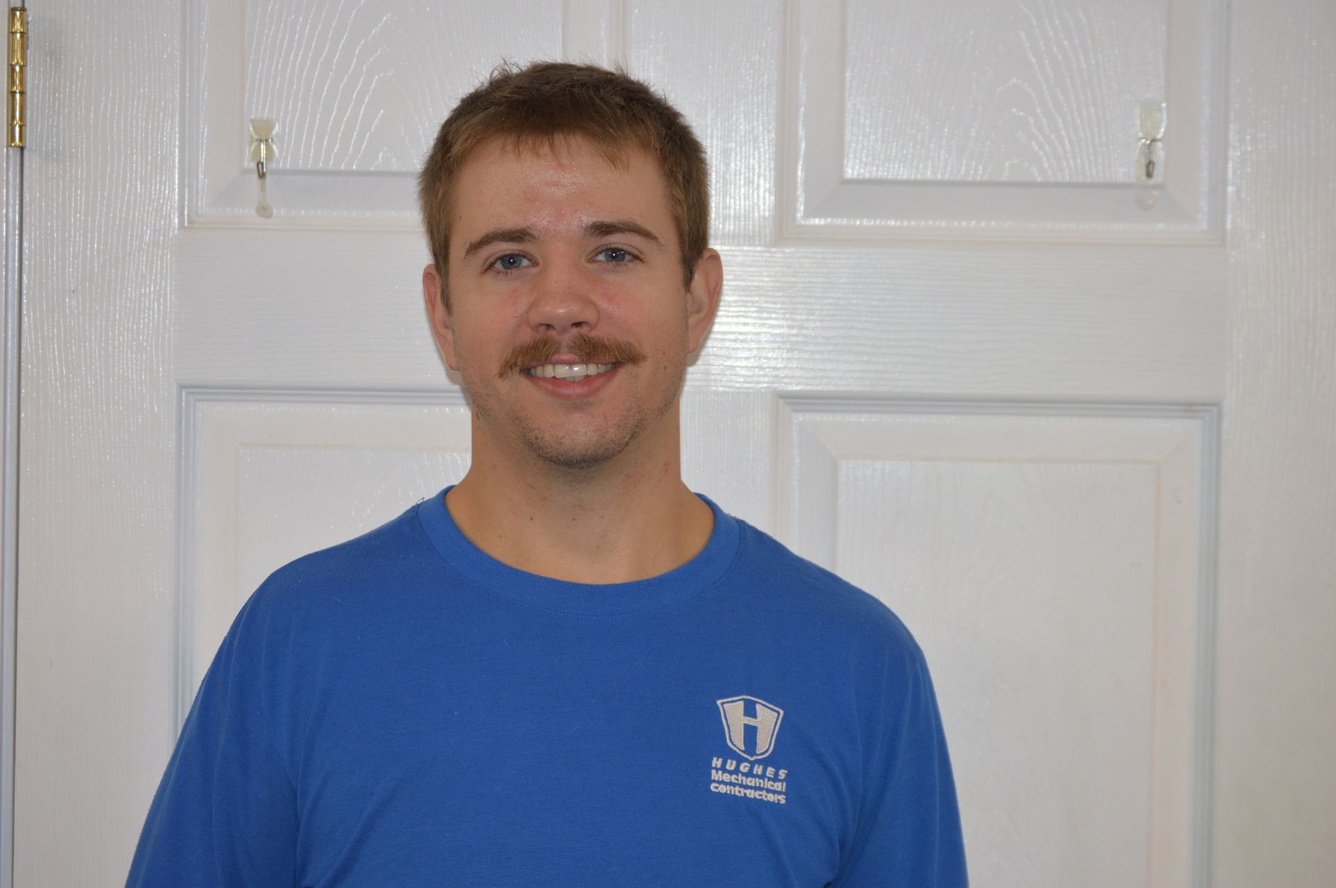 Man with a mustache wearing a blue shirt smiles in front of a white door.