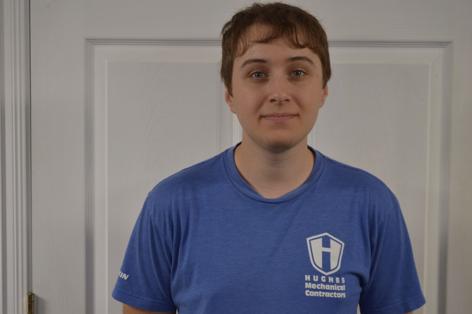 Person in blue shirt stands in front of a white door. The shirt has a logo.