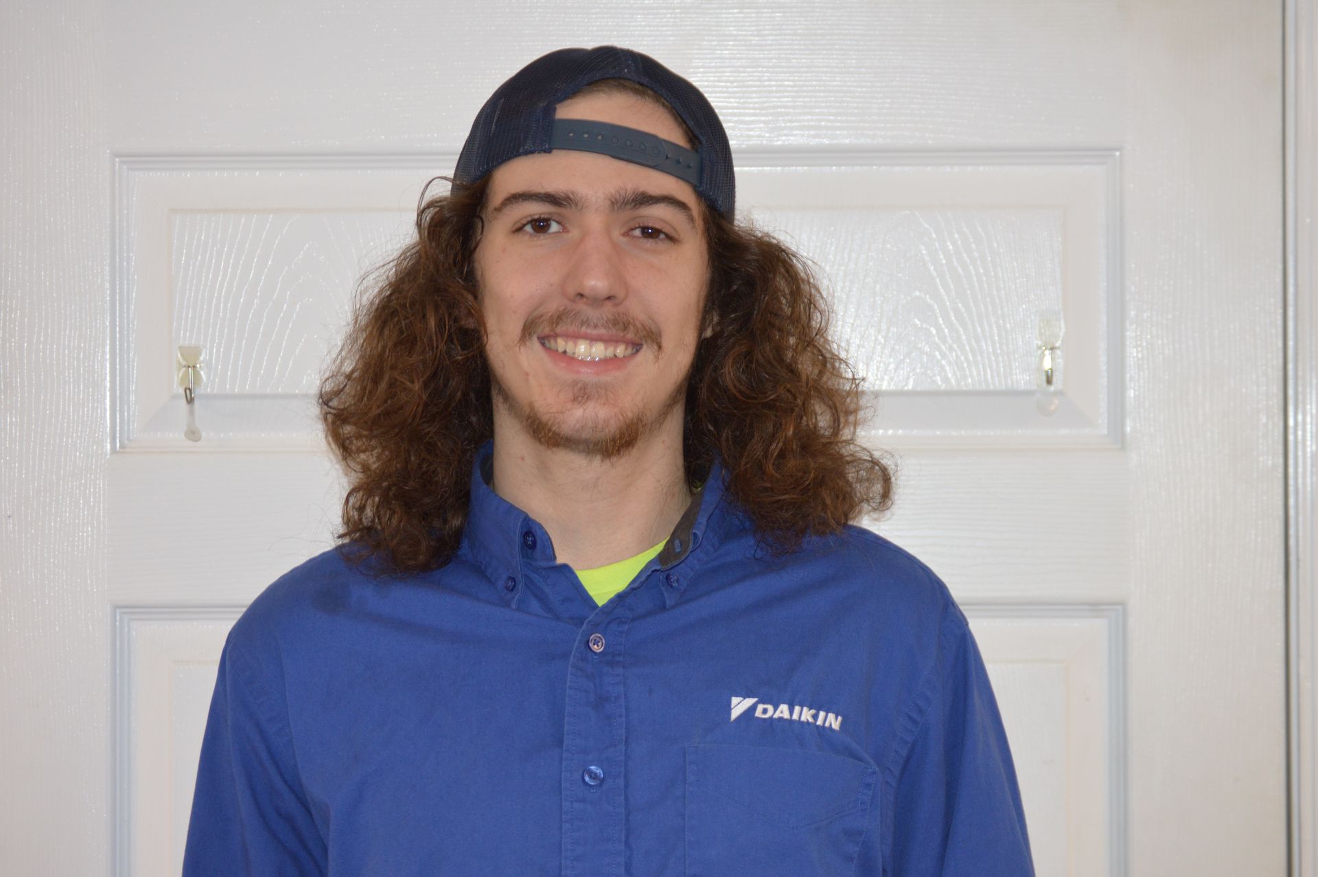 Man with long curly hair wearing a blue Daikin uniform and backwards baseball cap smiles in front of a white door.