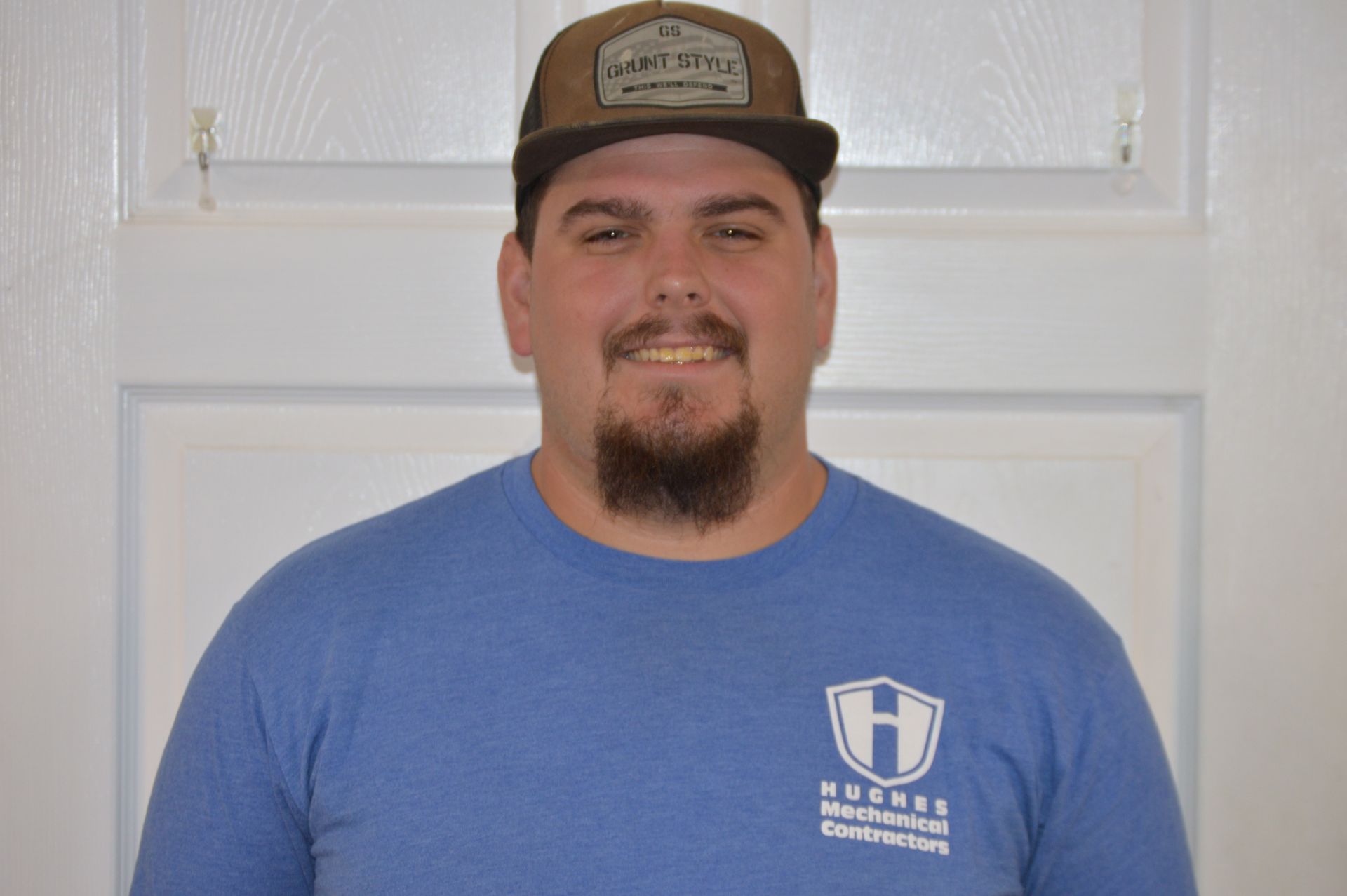 Man in blue shirt and baseball cap smiles at the camera, in front of a white door.