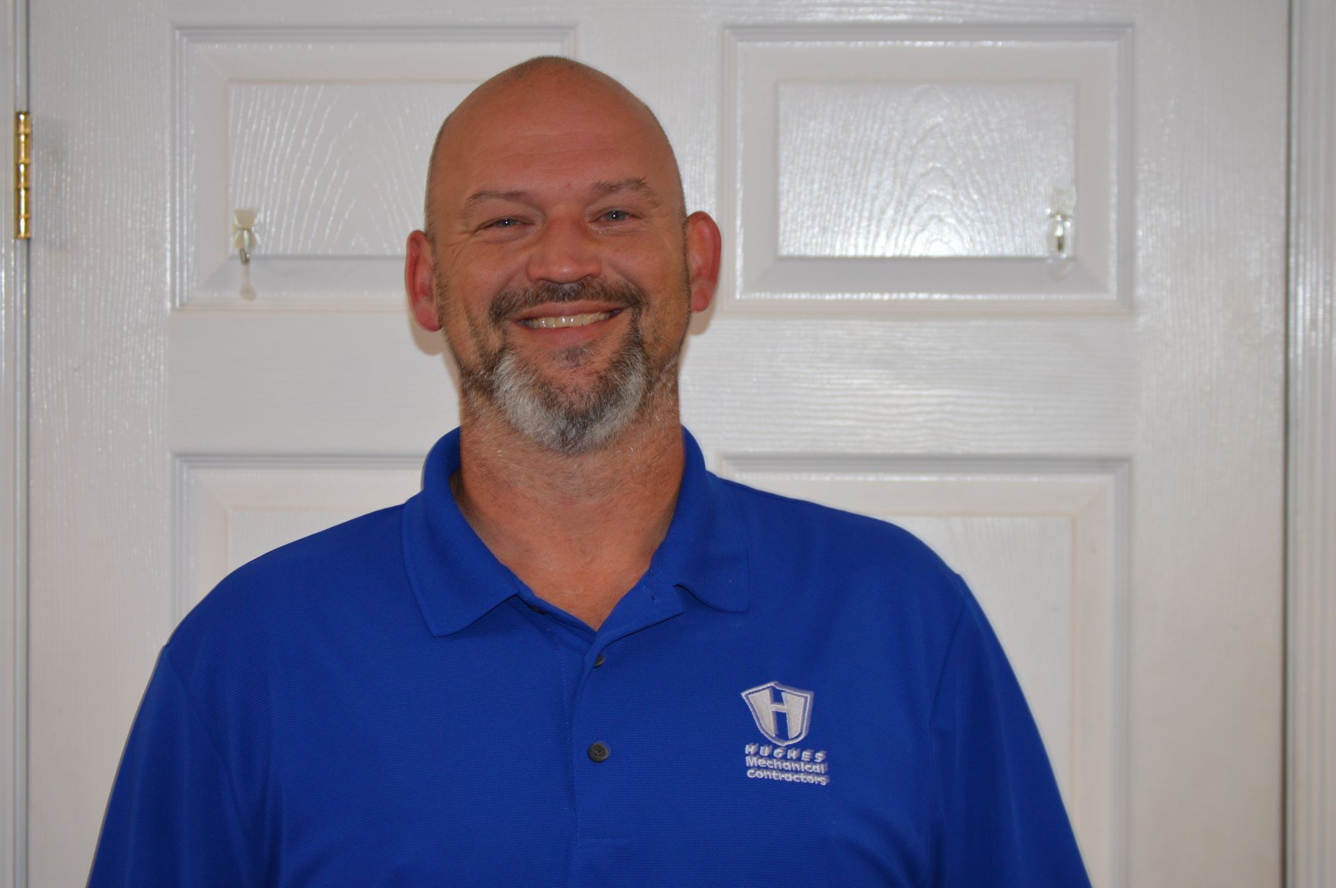Man with shaved head and beard in a blue polo shirt smiles in front of a white door.