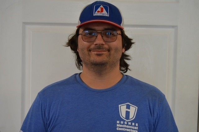 Man in blue hat and shirt, wearing glasses, smiles in front of a white door.