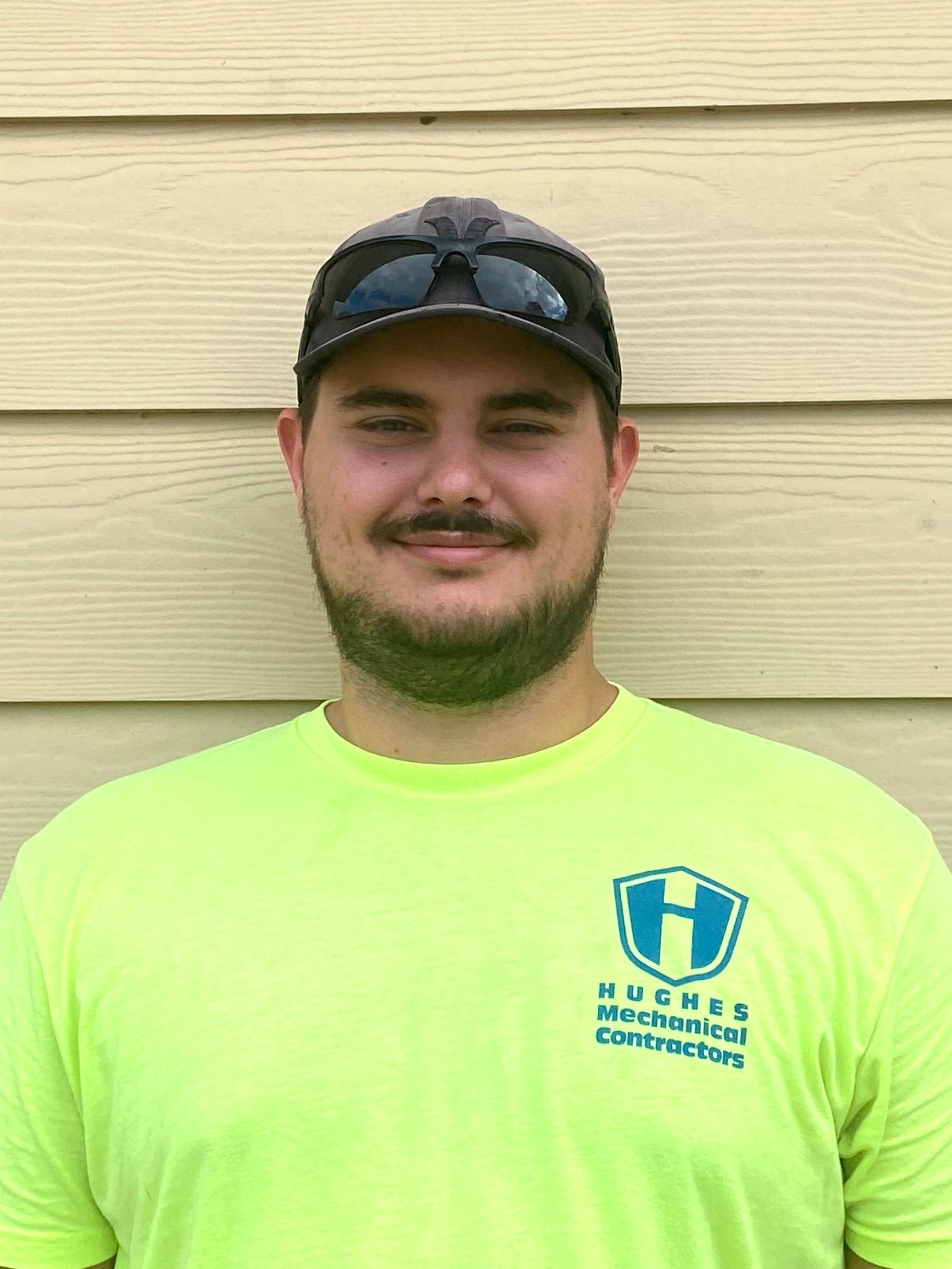 Man wearing a cap and bright yellow shirt, standing in front of a light brown wall.
