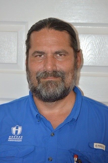 Man with long hair and beard in blue shirt, looking at the camera. A logo is visible on the shirt.