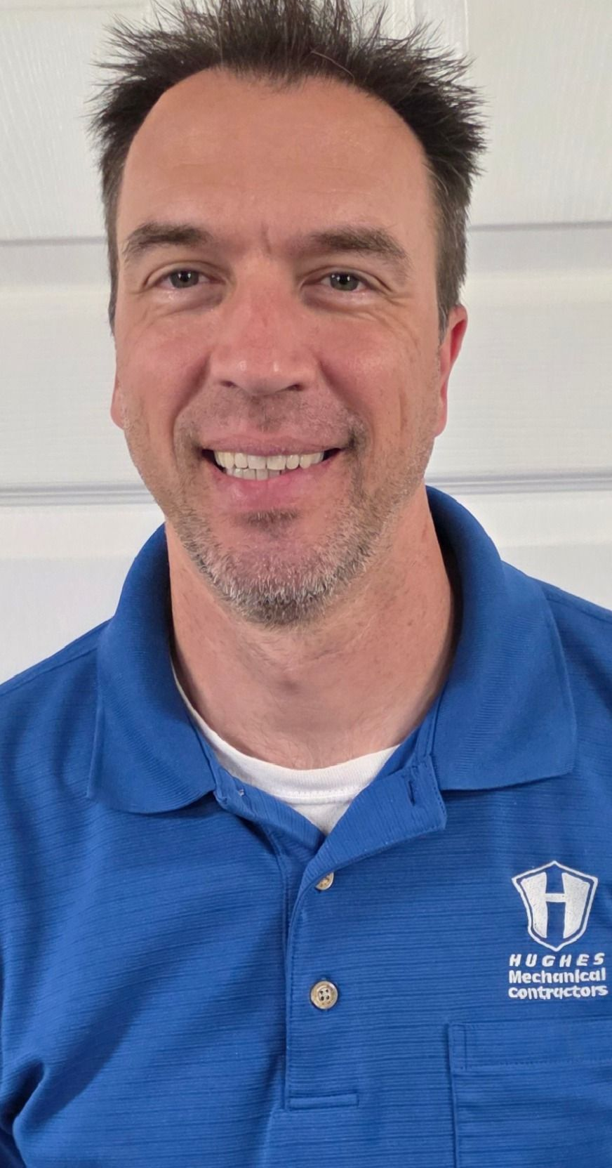 Man with short brown hair smiles, wearing a blue polo shirt, indoors.
