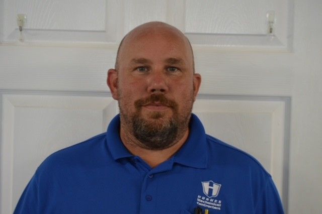 Bald man with beard wearing a blue polo shirt, standing in front of a white door.