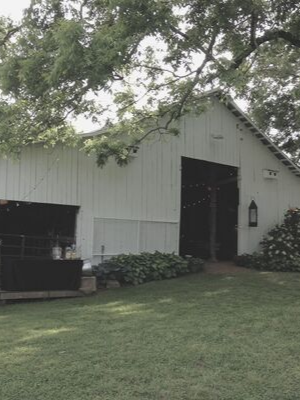 White barn with open doors and a dark horse trailer, shaded by trees on a grassy lawn.
