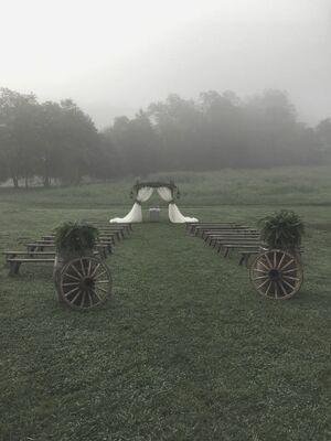 Foggy outdoor wedding setup with chairs lining a grassy aisle toward a white arch and gazebo