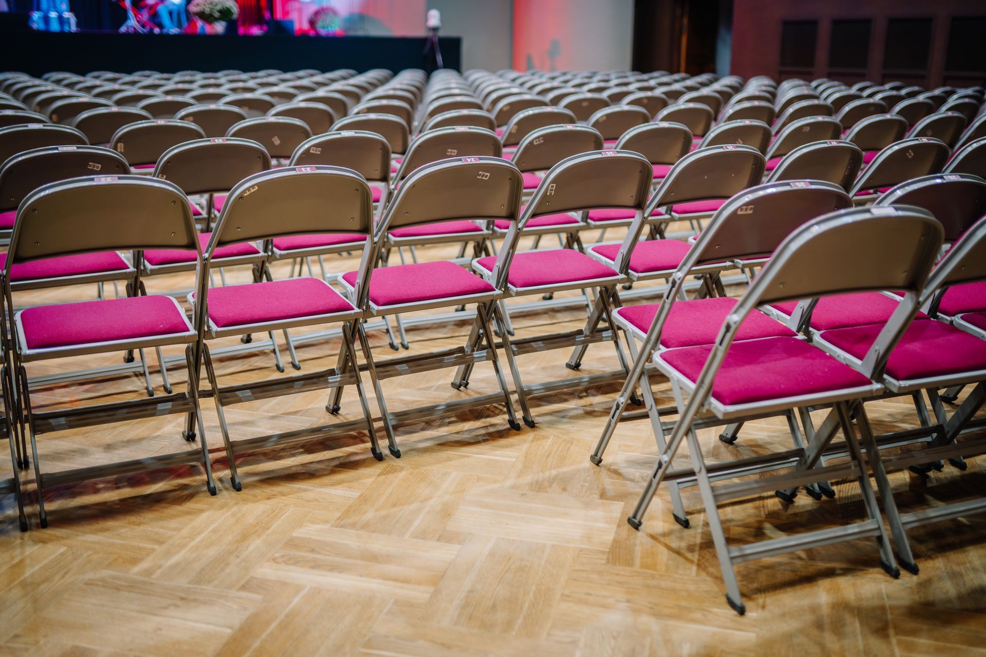 Rows of empty folding chairs with pink seats face a stage in a large room.
