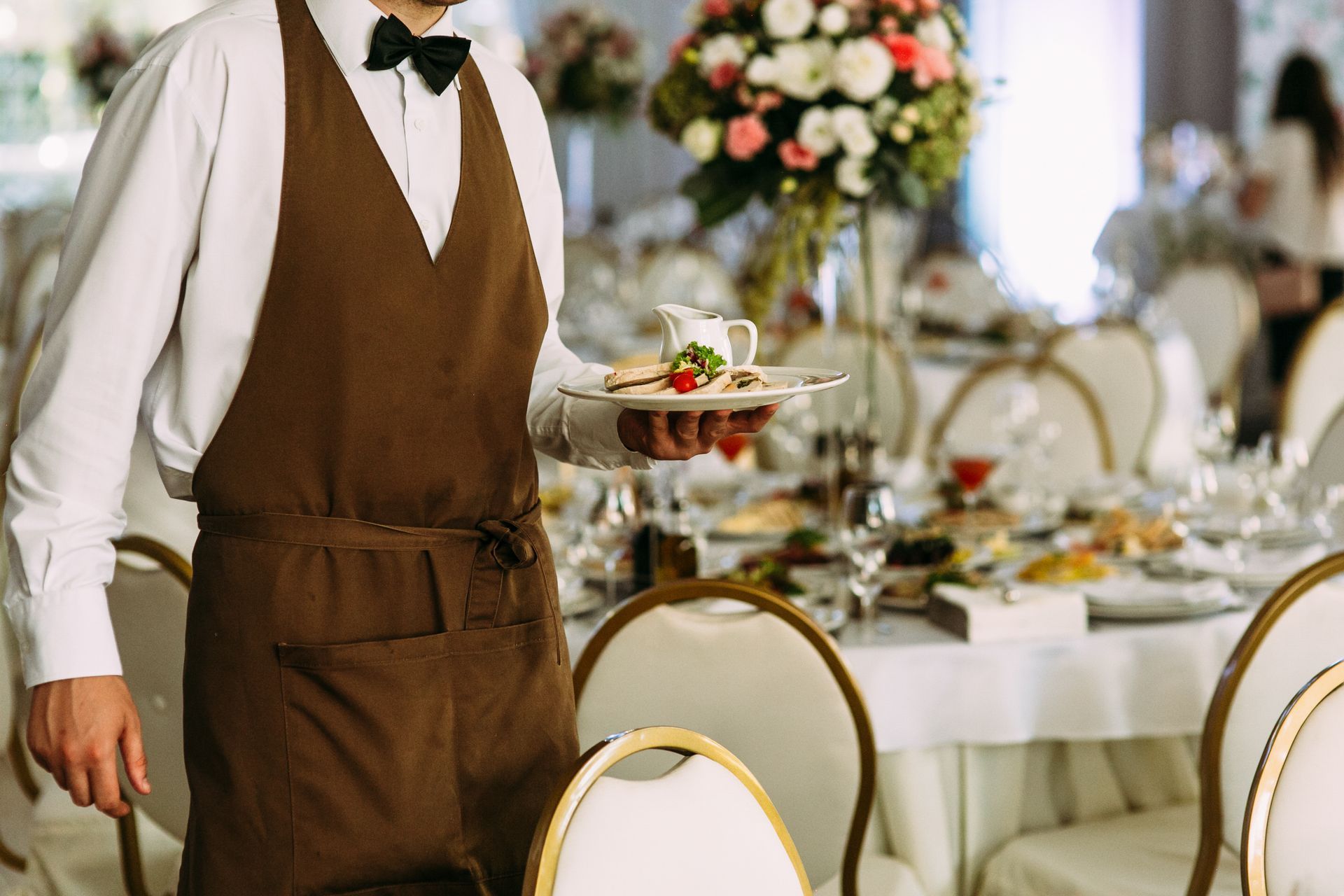 Waiter in brown apron holds plate of food near a banquet table set for a formal event.