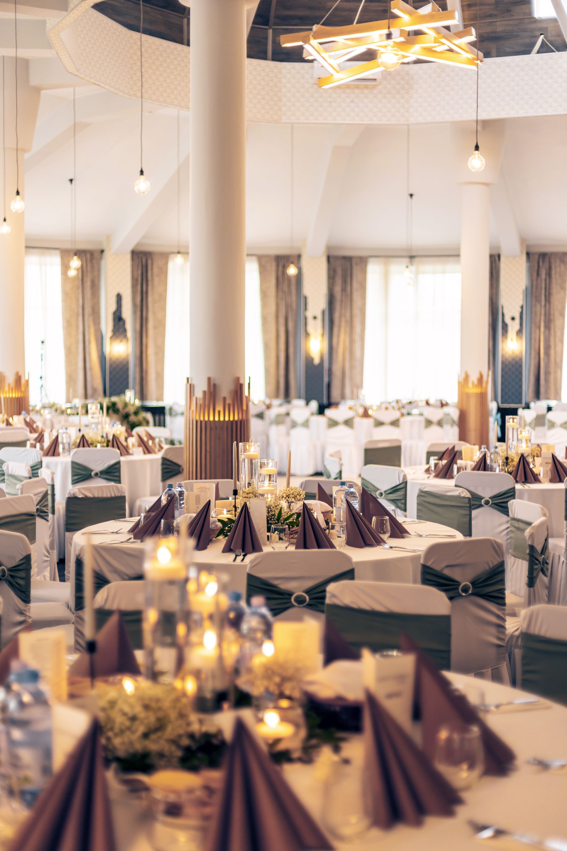 Wedding reception hall with round tables, white linens, and burgundy napkins. Green chair sashes, candles, and chandeliers.