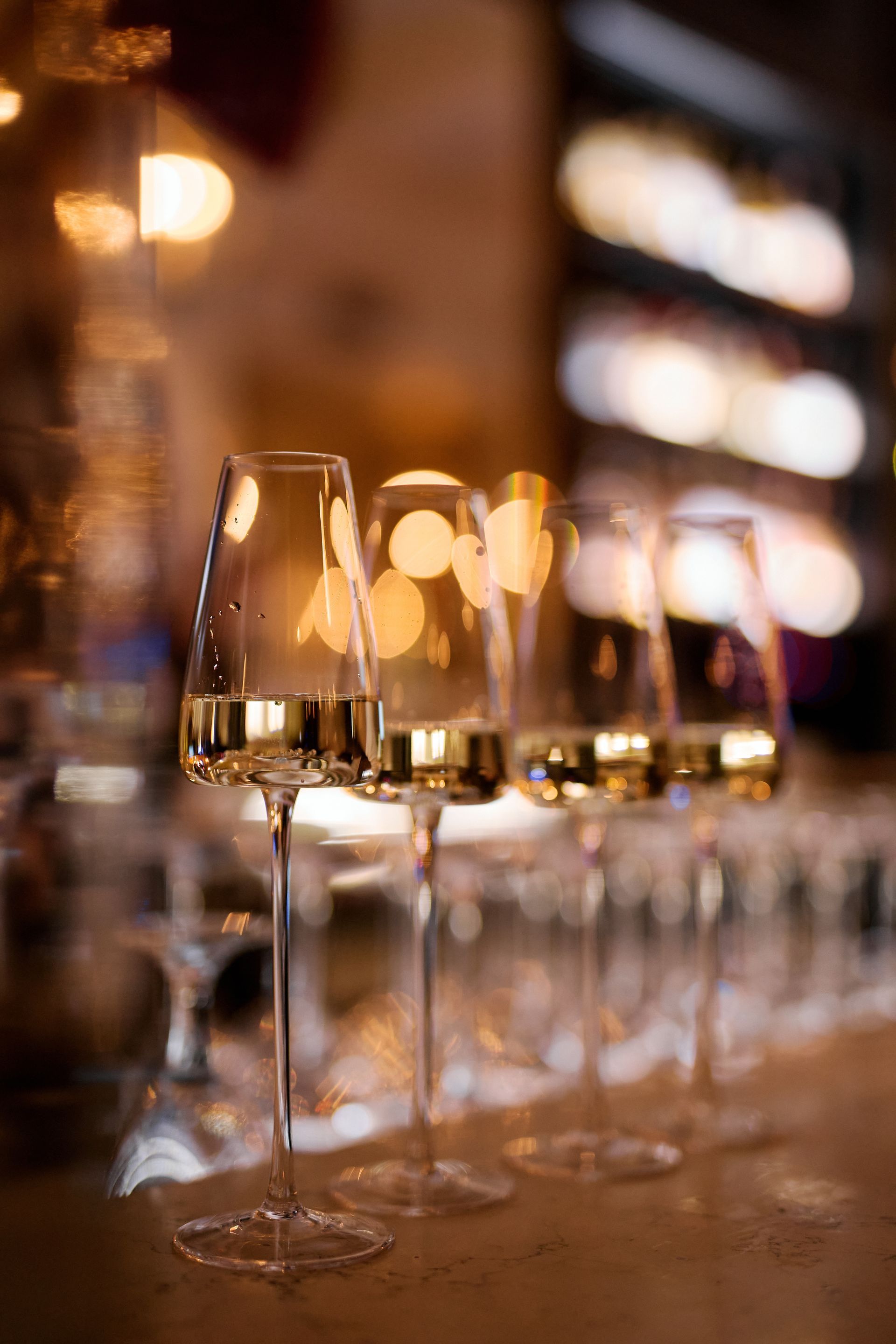 Five champagne flutes filled with light-colored liquid on a bar, bokeh lights in the background.