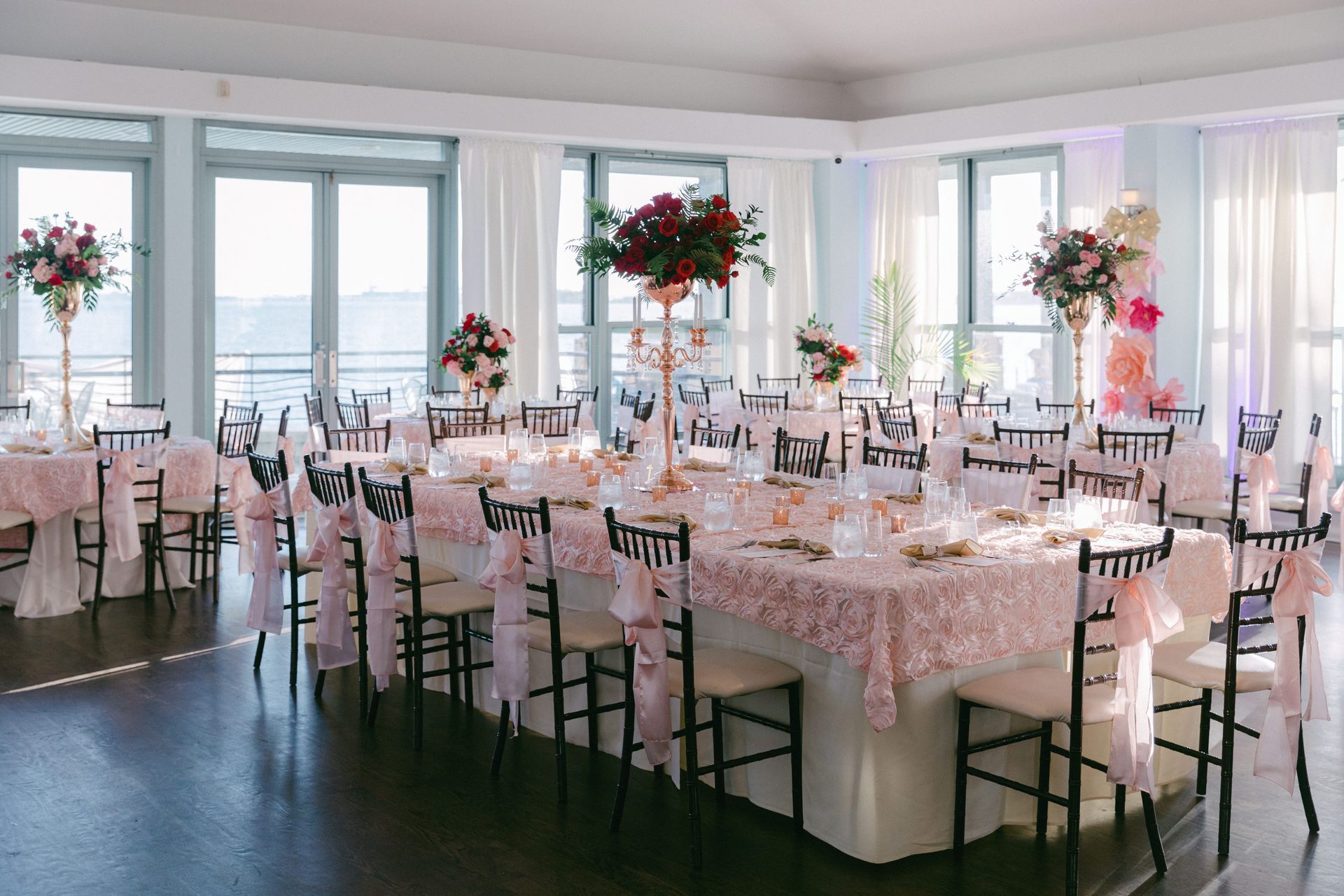Elegant event space set for a wedding; tables with pink floral centerpieces, black chairs, and large windows.