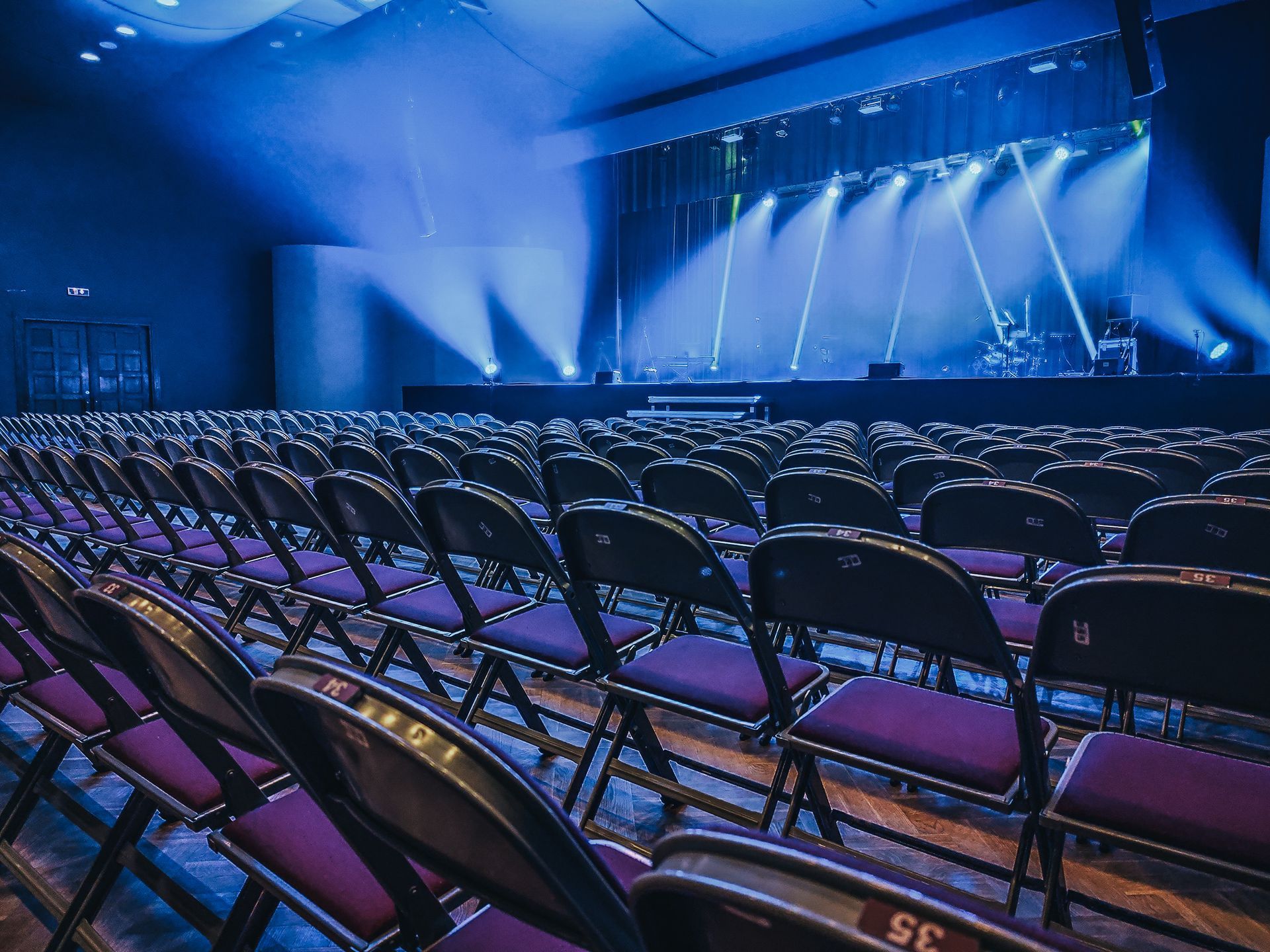 Empty auditorium with rows of purple-seated chairs facing a stage lit with blue lights.