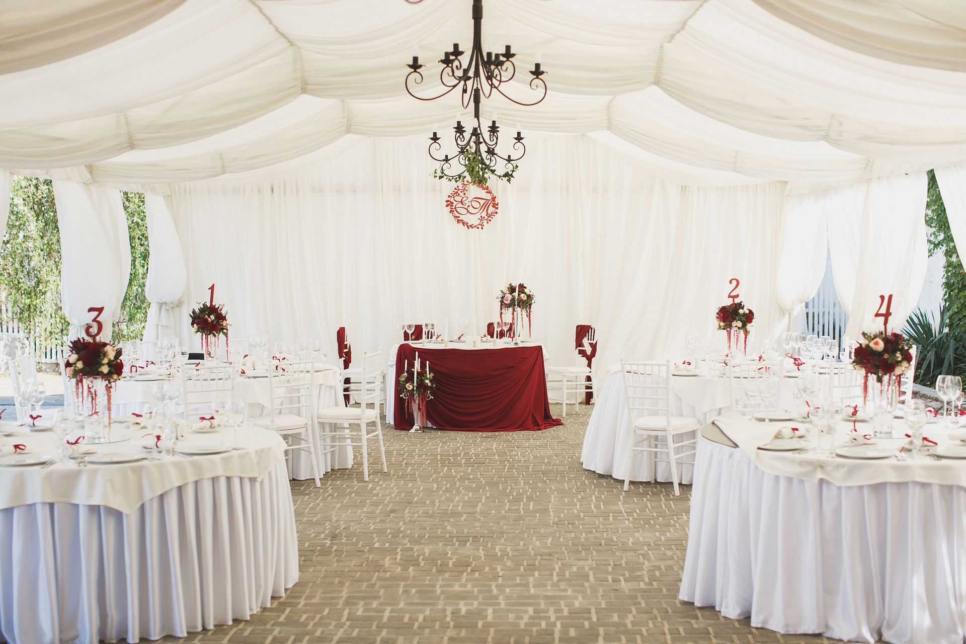 White tent decorated for a wedding reception with round tables, red and white floral centerpieces, and a dark red head table.