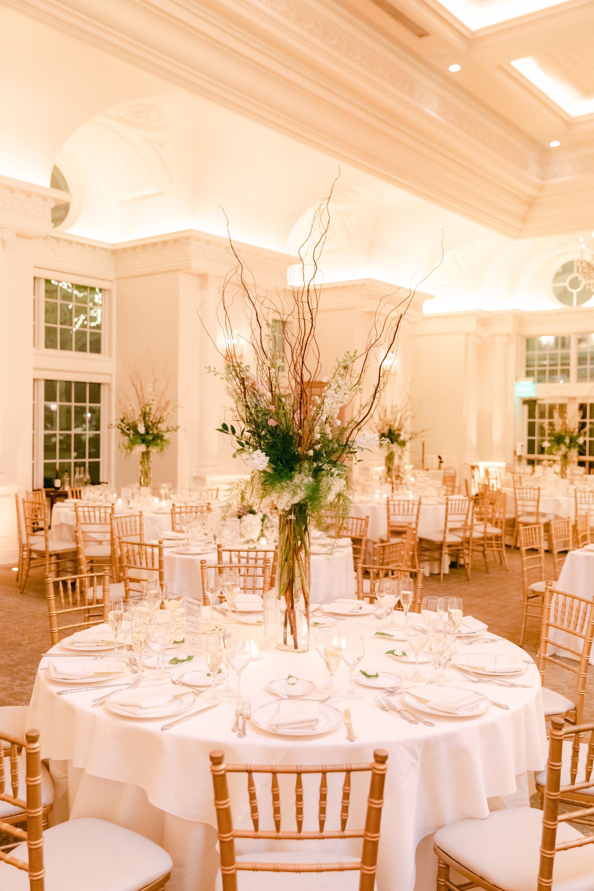 Elegant wedding reception with round tables, floral centerpieces, and gold chairs in a bright, white room.