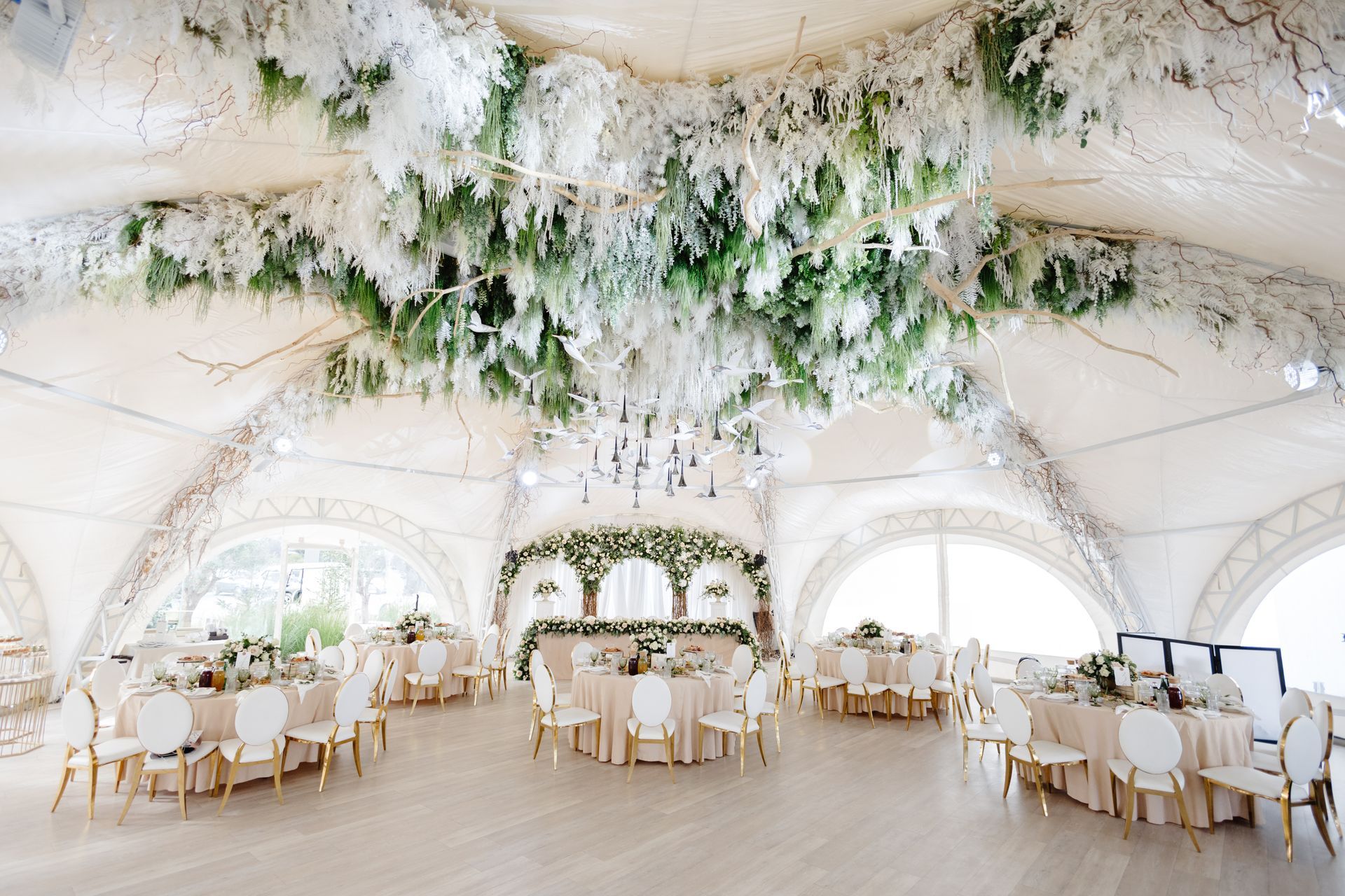 Elegant wedding reception in a tent, decorated with white flowers, round tables, and arched windows.