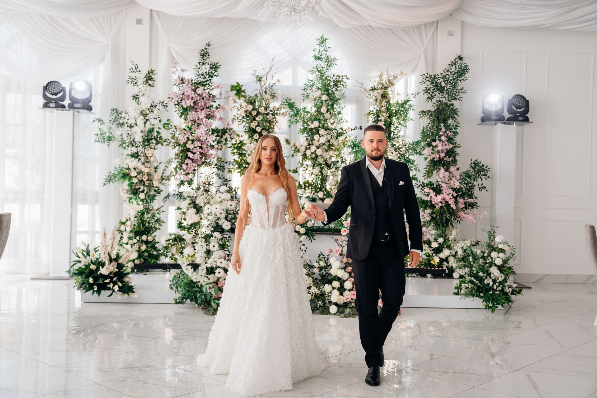 Bride and groom holding hands, walking towards the camera. Floral backdrop in white and green. White and black outfits.