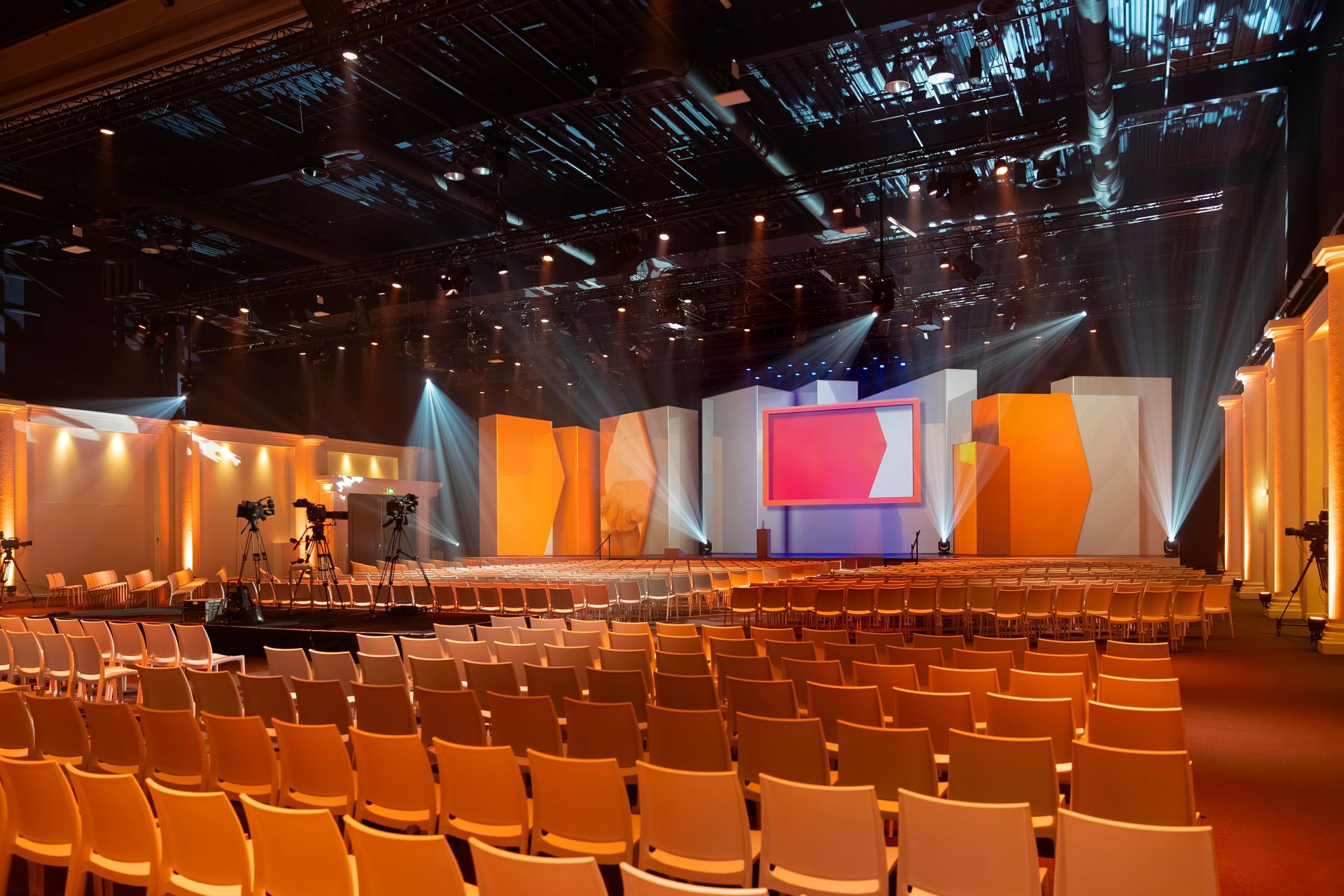 Empty auditorium with orange chairs facing a stage. Stage has a large screen and orange/white backdrop.