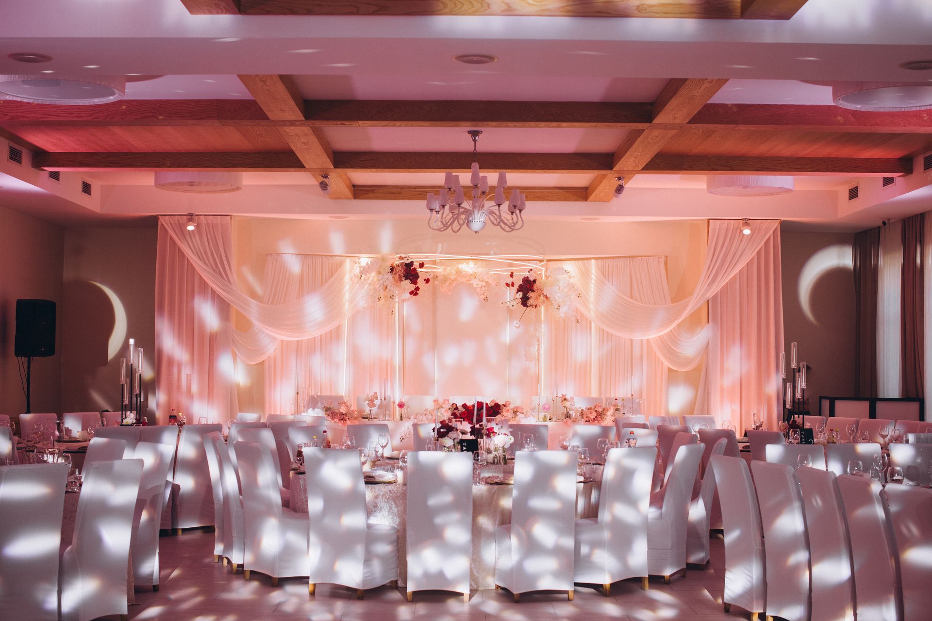 Wedding reception hall, decorated with white chairs, draping, and soft pink lighting.