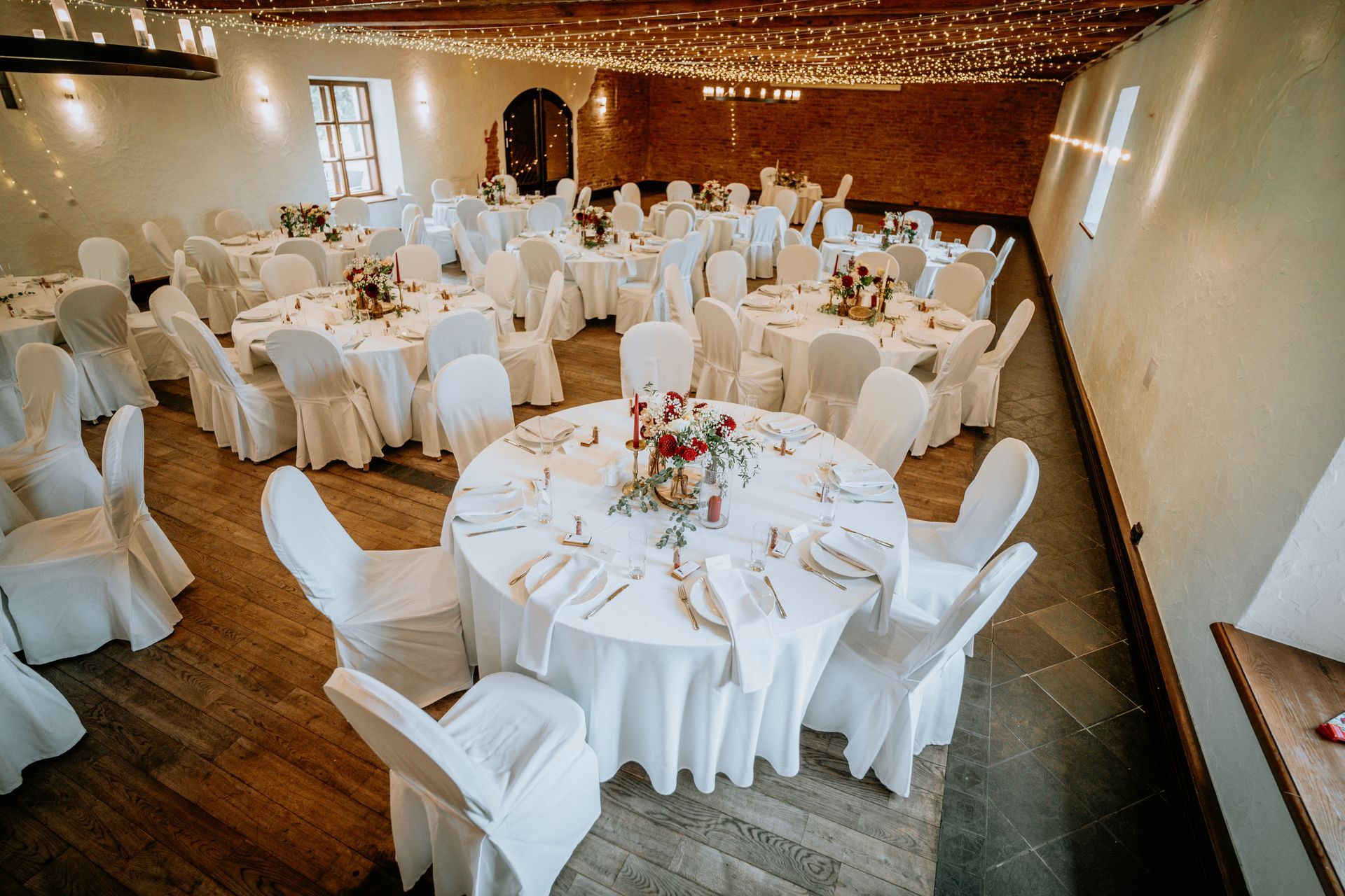 Wedding reception hall with round tables set for guests, floral centerpieces, white linens, and string lights.