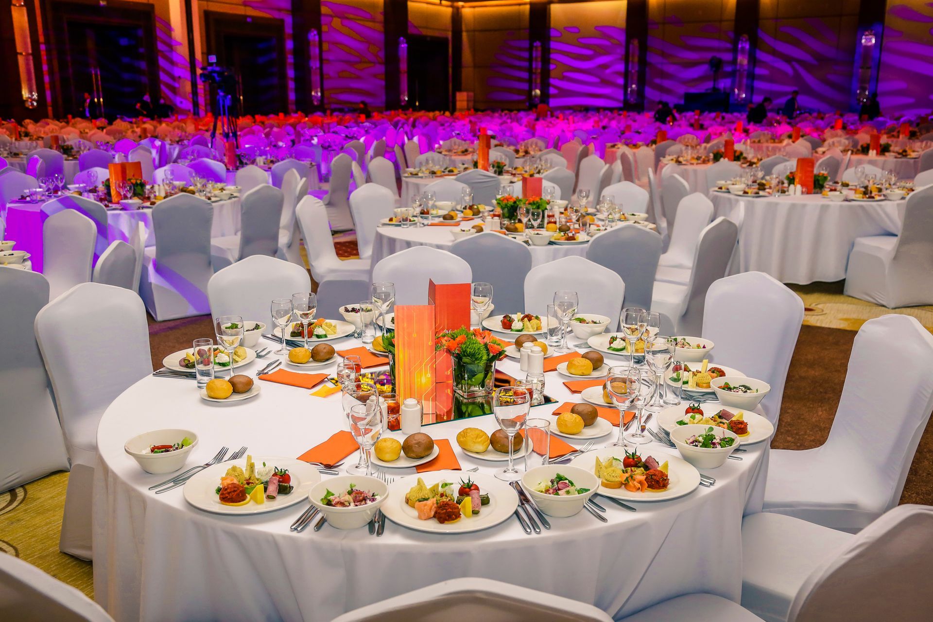 Round tables set for a banquet in a brightly lit hall. Each table has food, centerpieces, and white chair covers.
