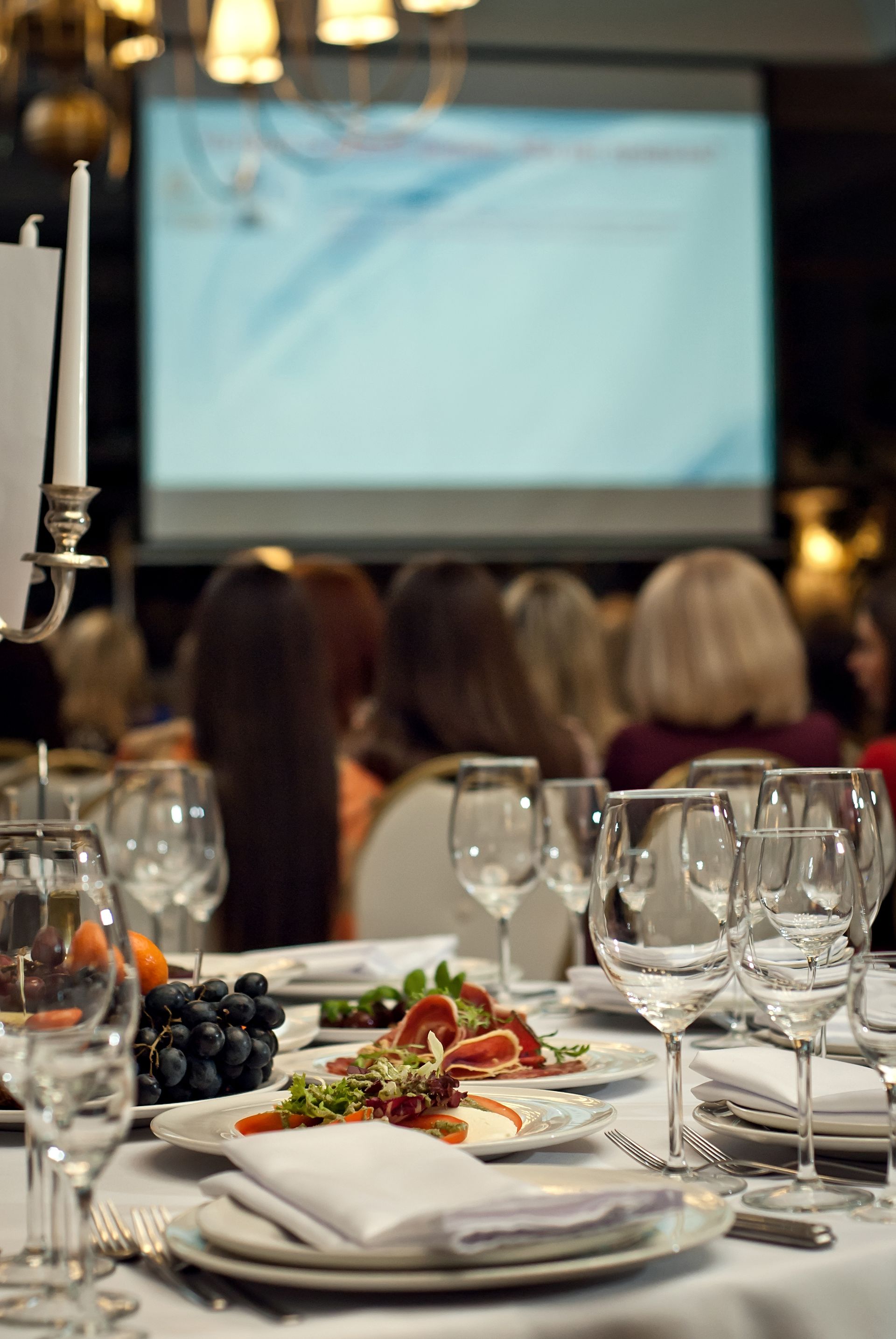 Table setting at an event, plates of food, wine glasses, audience in the background, screen displaying visuals.