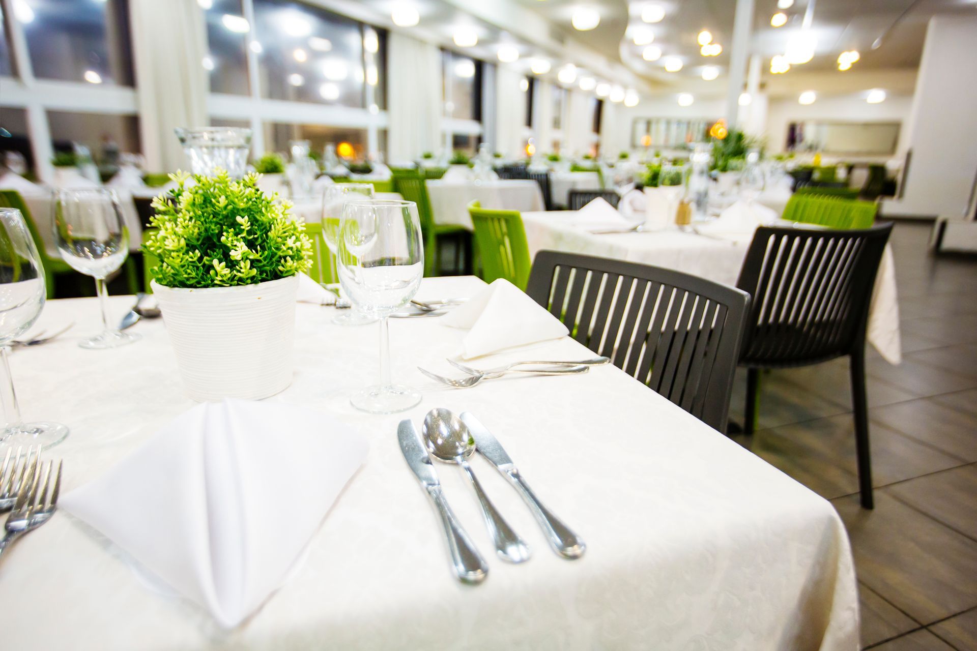 Restaurant dining room with white tablecloths, silverware, green chairs, and large windows.