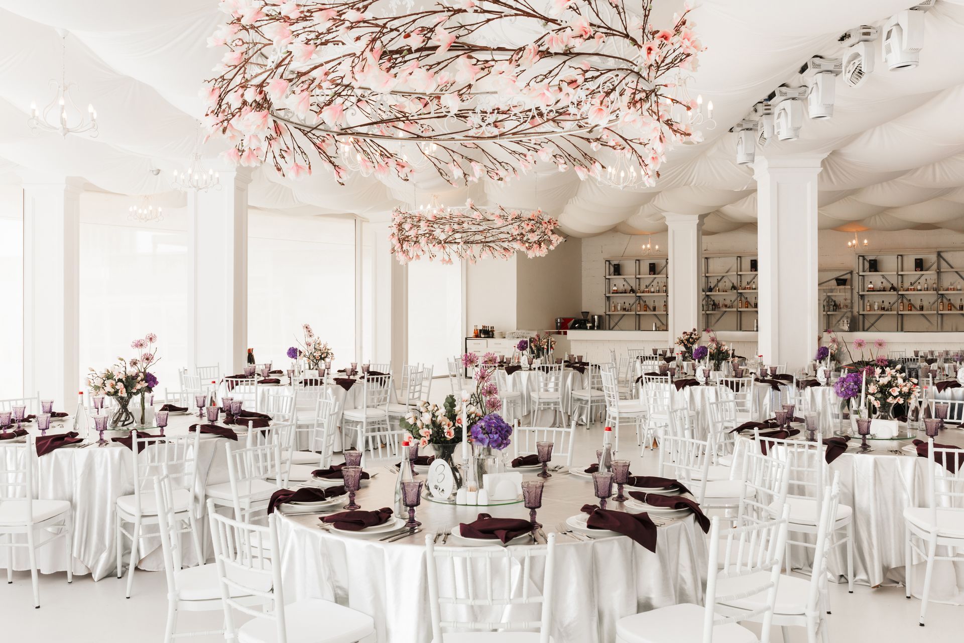 Elegant white wedding reception with round tables, floral centerpieces, and cherry blossom ceiling decor.