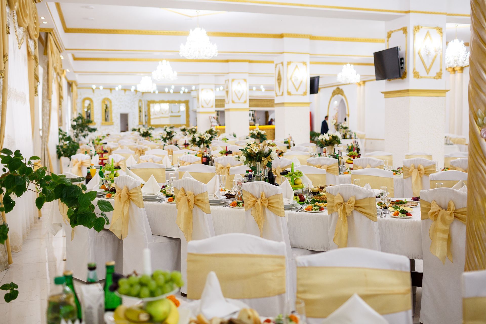 Elegant banquet hall with decorated tables, chairs, and floral centerpieces; white and gold decor.
