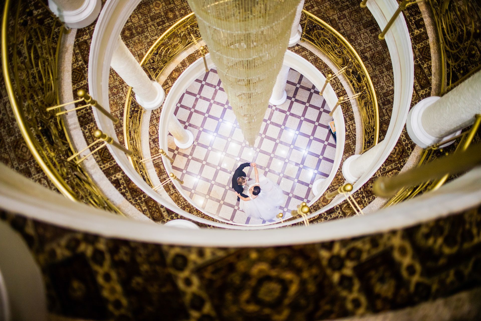 Bride and groom stand at the bottom of a circular staircase, viewed from above, ornate gold and white decor.