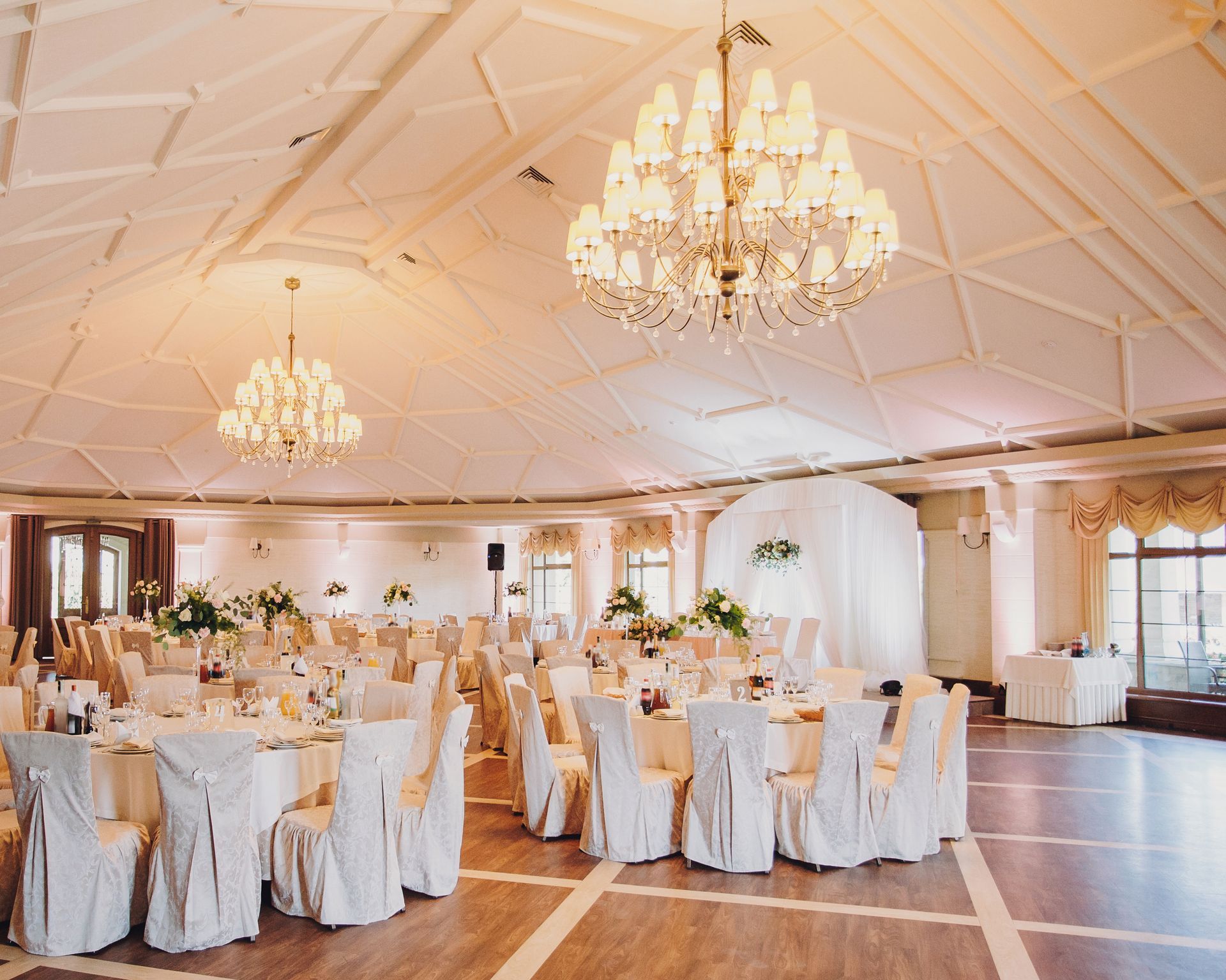 Elegant ballroom set for a wedding, with round tables, white chairs, and large chandeliers.