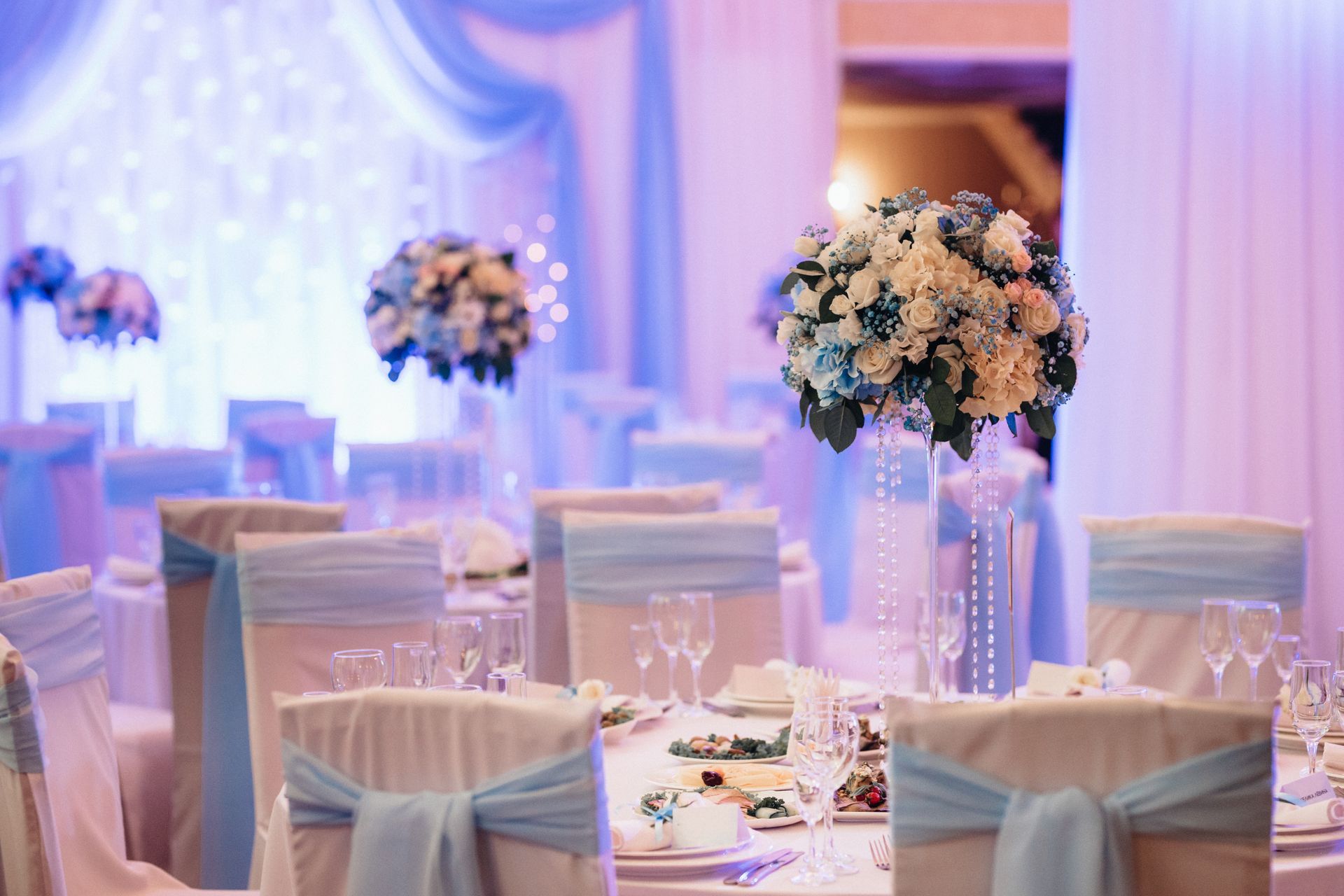Elegant event hall decorated in light blue, with floral centerpieces on tables set for a celebration.