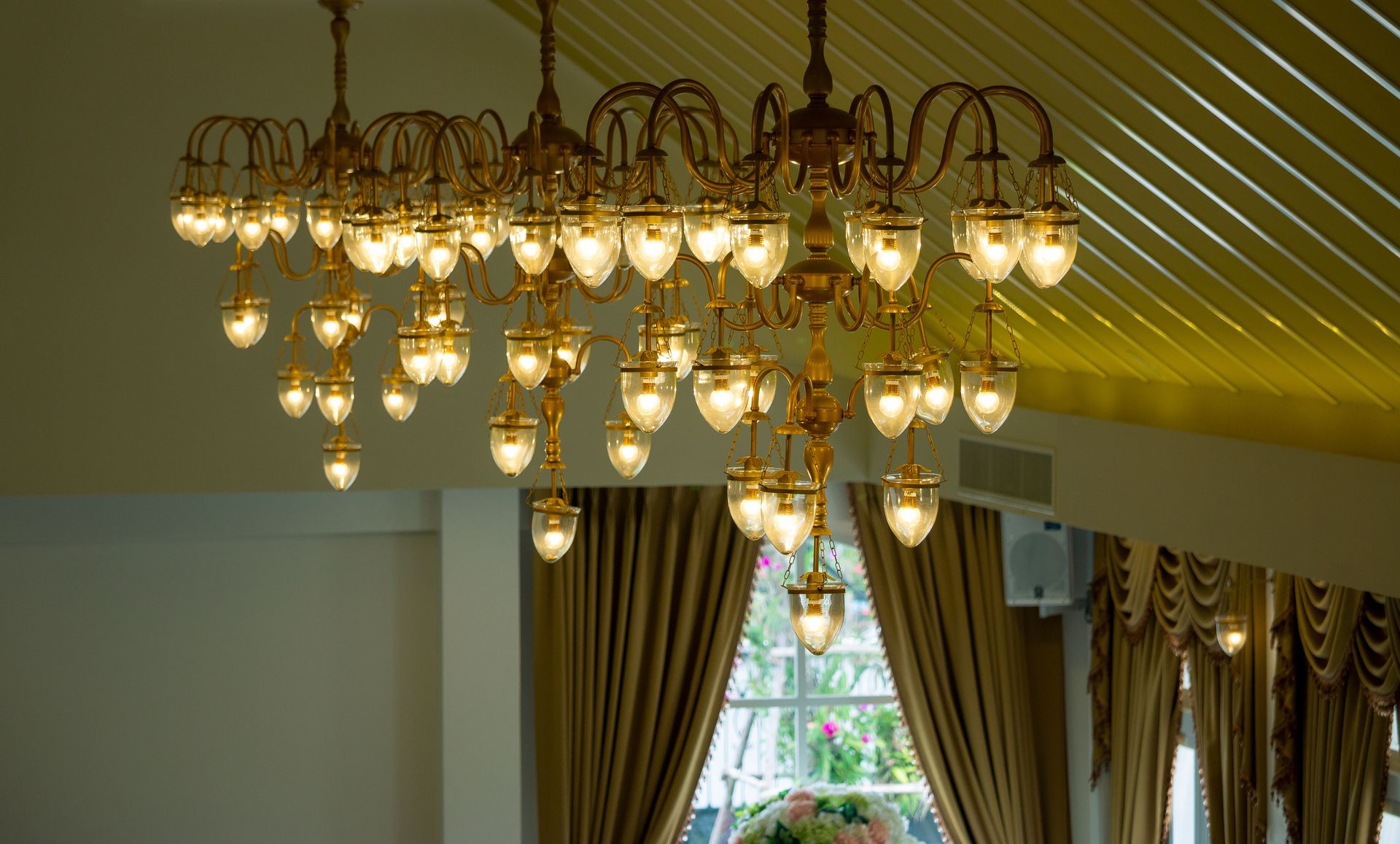 Golden chandelier with many lights, hanging in a room with a window and gold curtains.