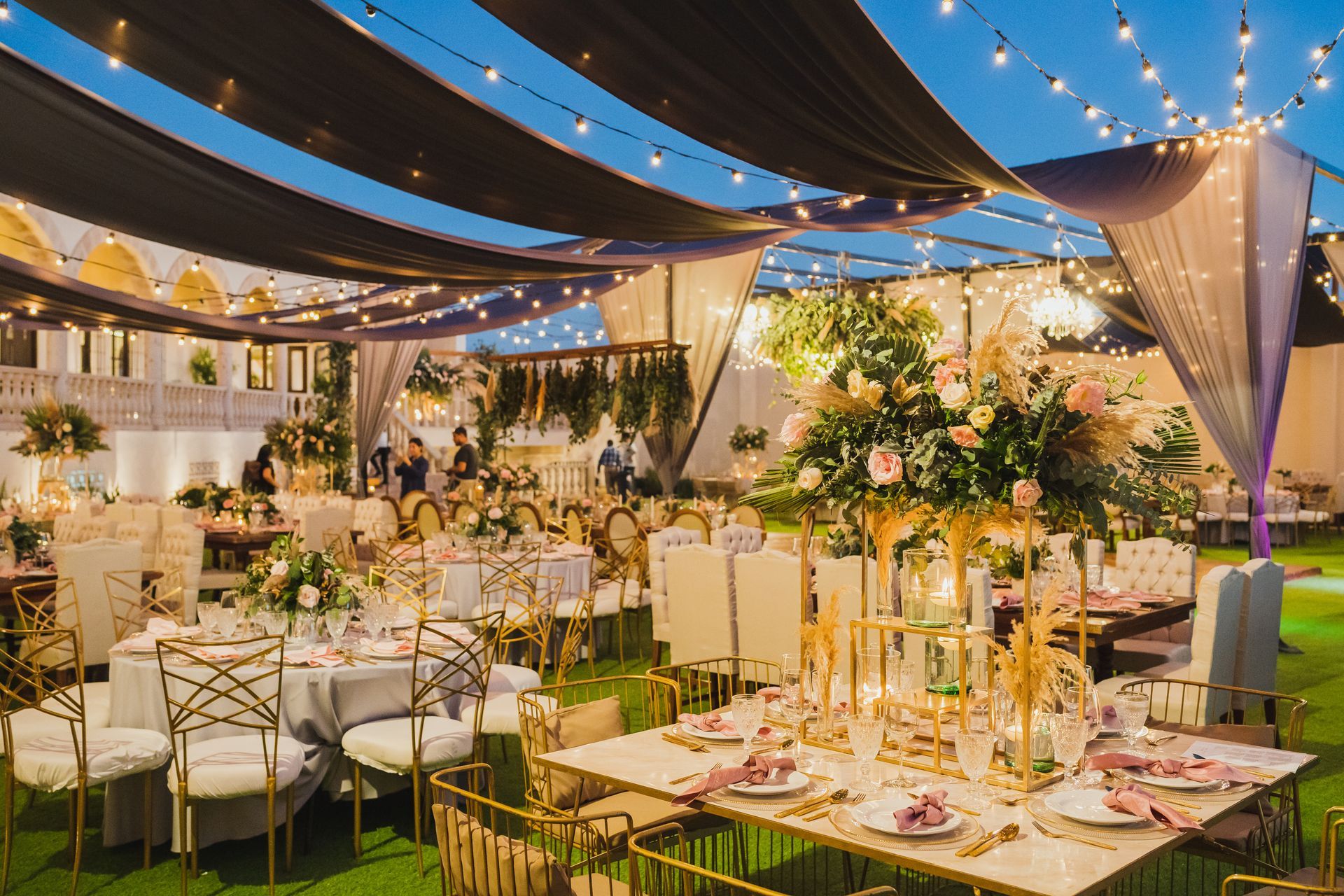 Outdoor wedding reception with decorated tables, string lights, and draping fabric.