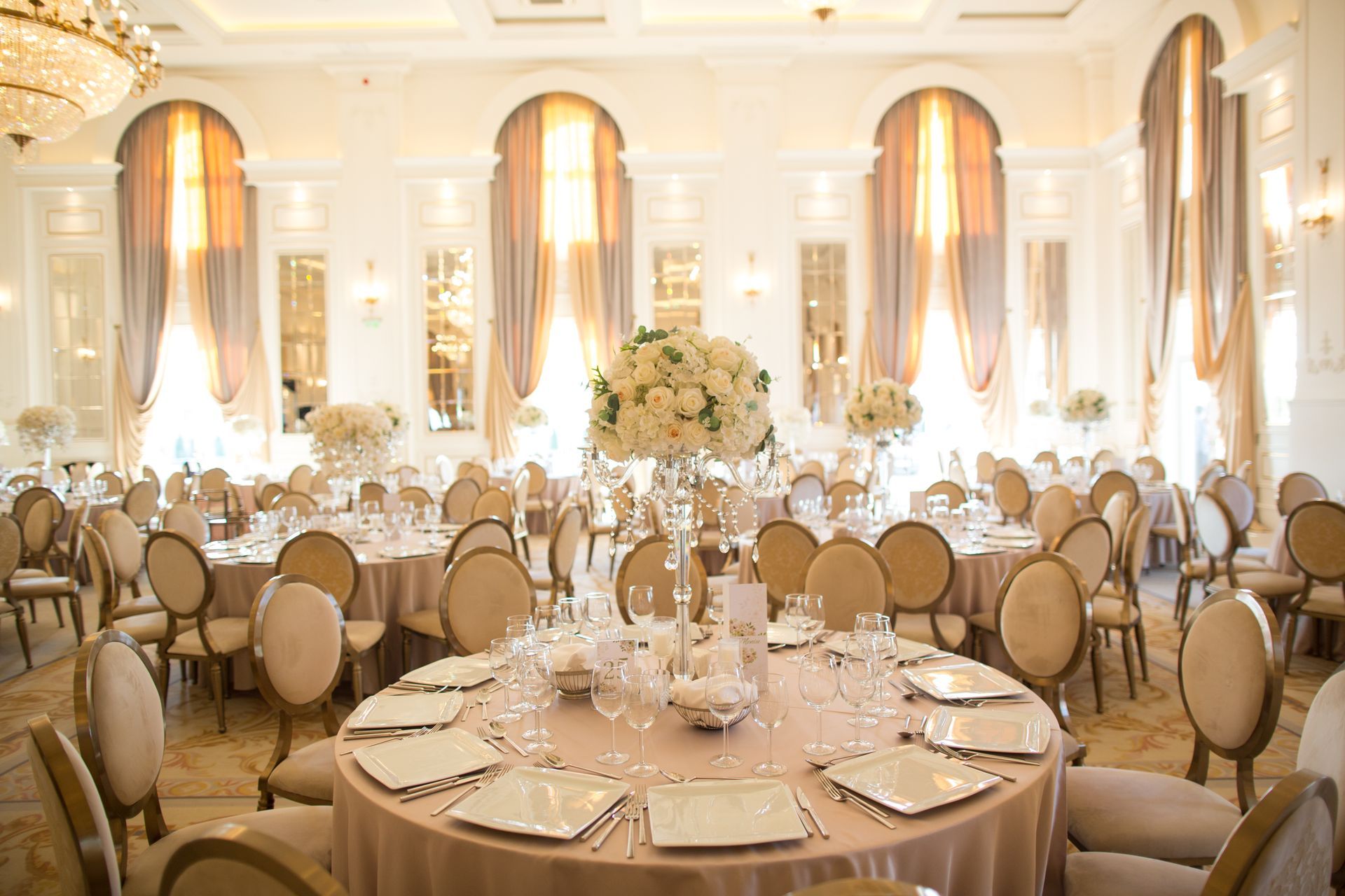 Elegant ballroom set for a formal event with round tables, floral centerpieces, and draped curtains.