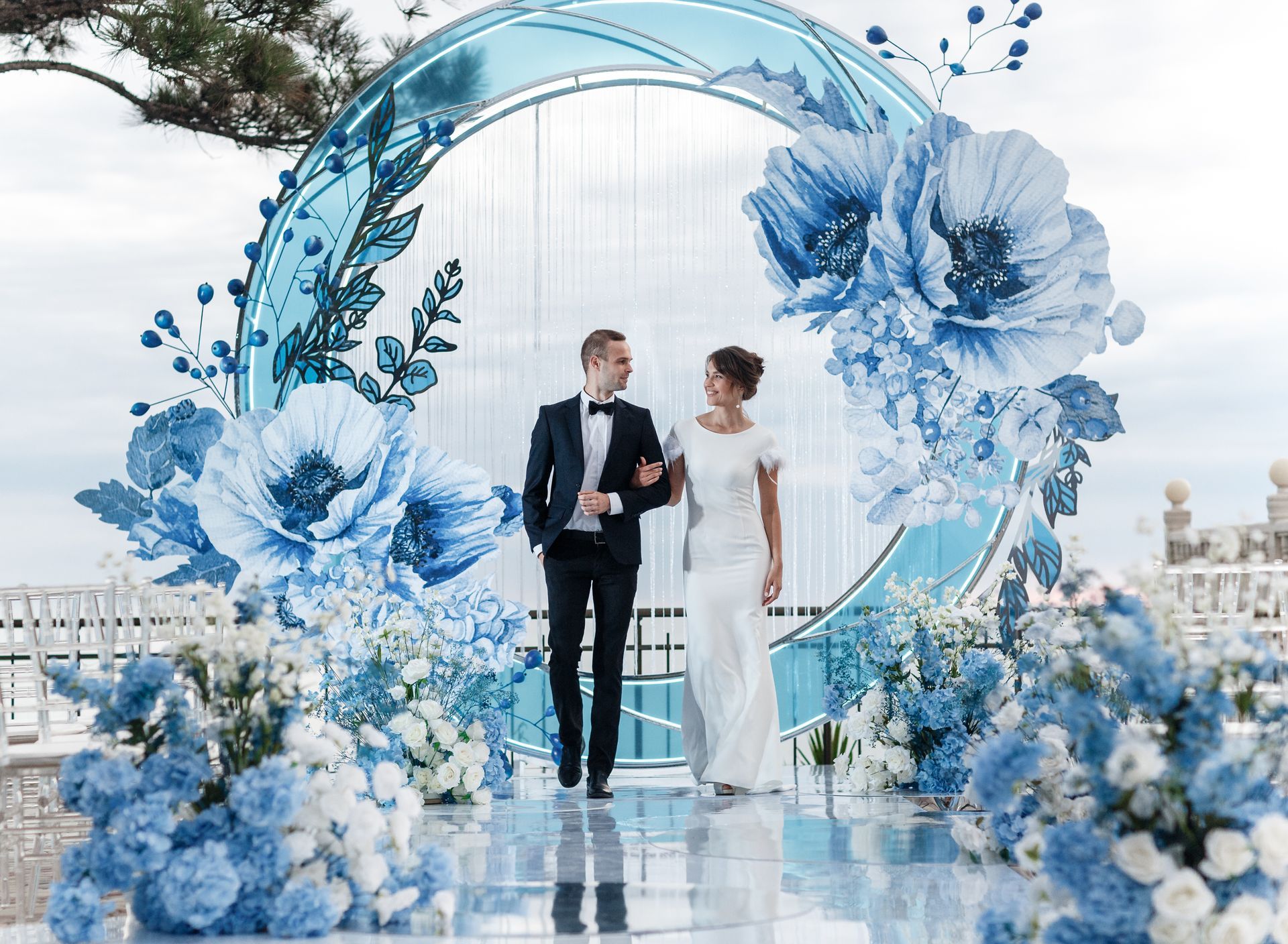 Couple walking under blue floral archway at an outdoor wedding.