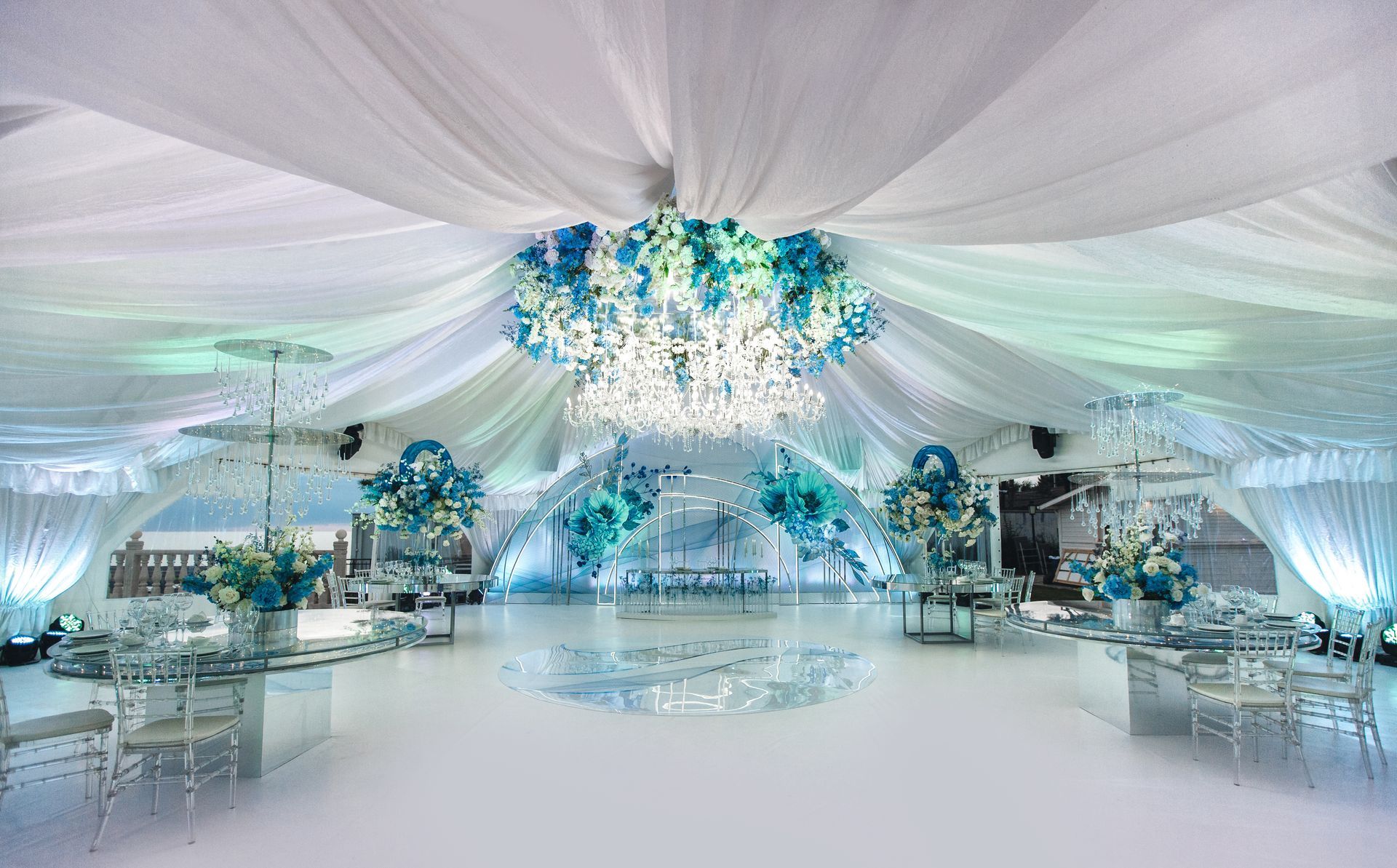 Elegant event space with white draping, a chandelier, and blue floral accents.
