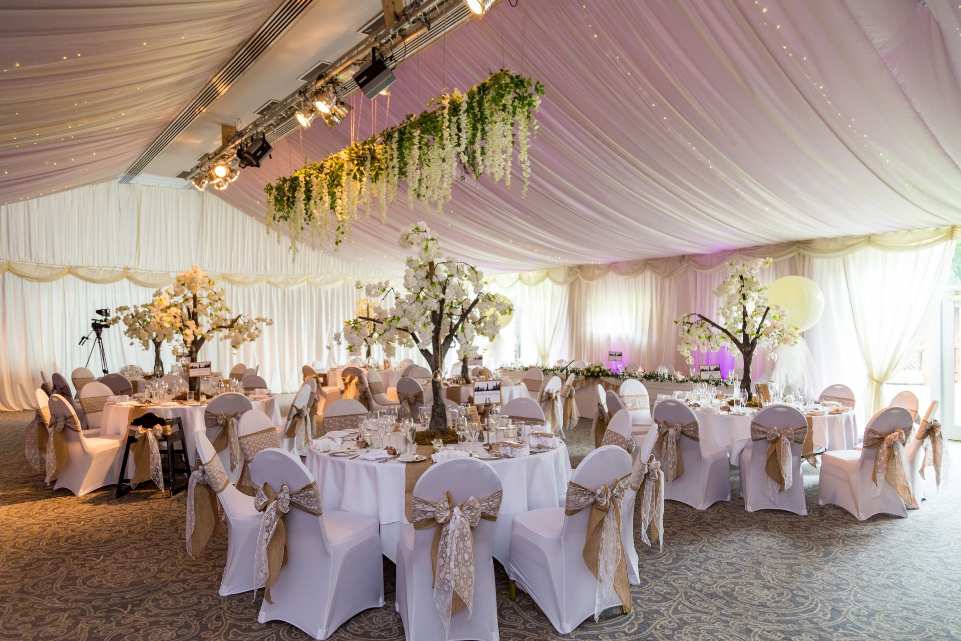 Elegant wedding reception in a white tent with round tables, floral centerpieces, and draped fabric.