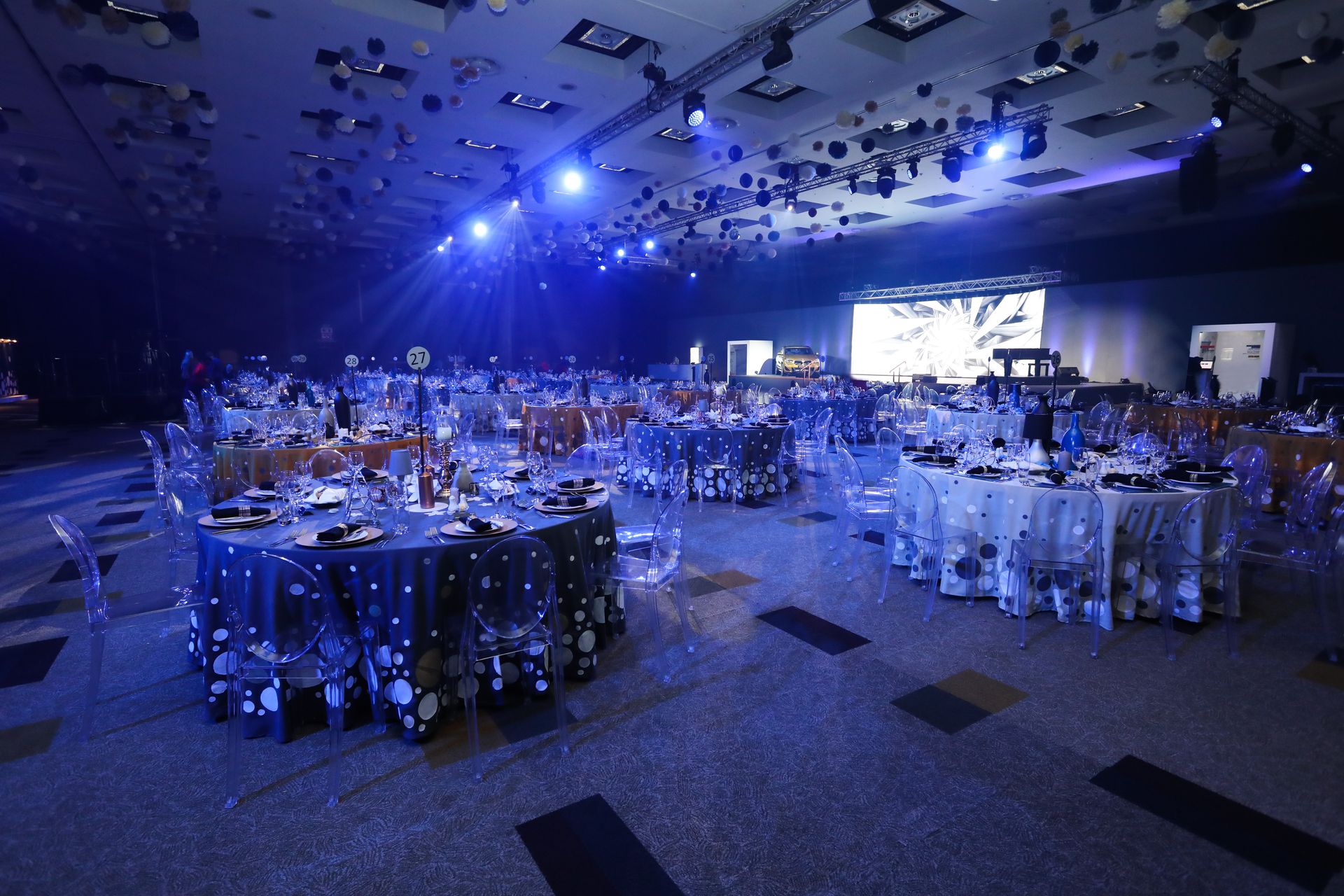 Large ballroom set for an event; round tables with tablecloths, blue and gold lighting.