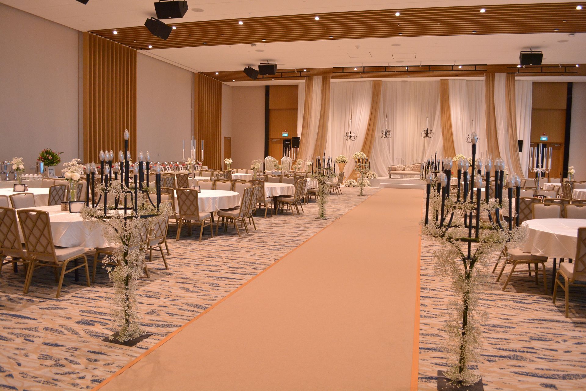 Elegant wedding reception setup; rows of tables, decorated stage, floral aisle runner, neutral tones.