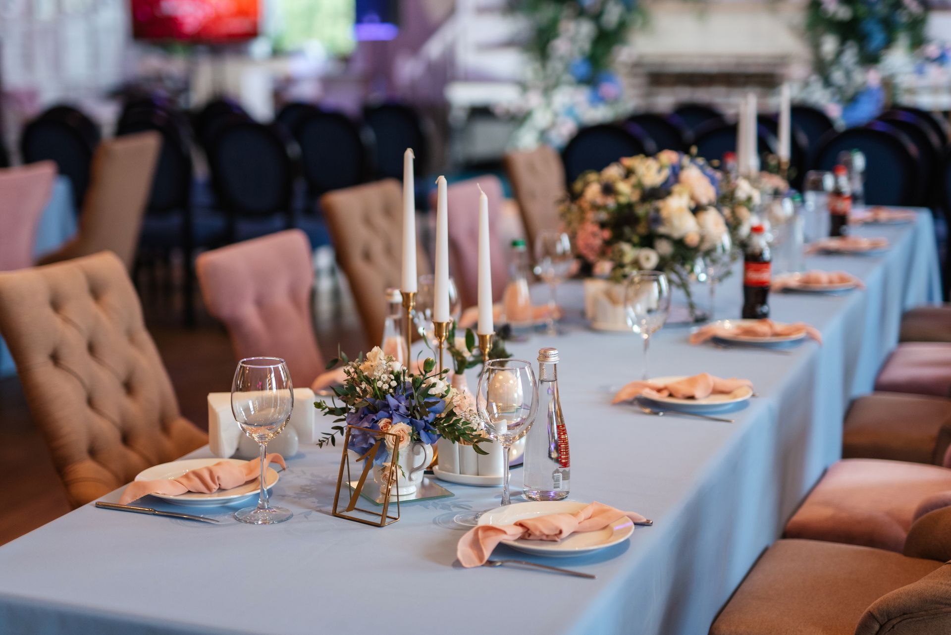 Wedding reception table set with blue tablecloth, floral arrangements, candles, and pink napkins.